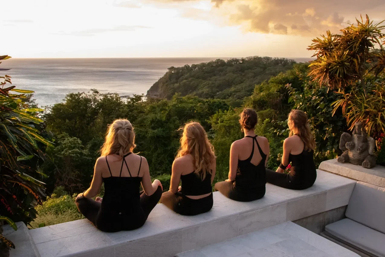Four women sitting cross-legged on a ledge, overlooking a lush green forest and ocean during sunset.