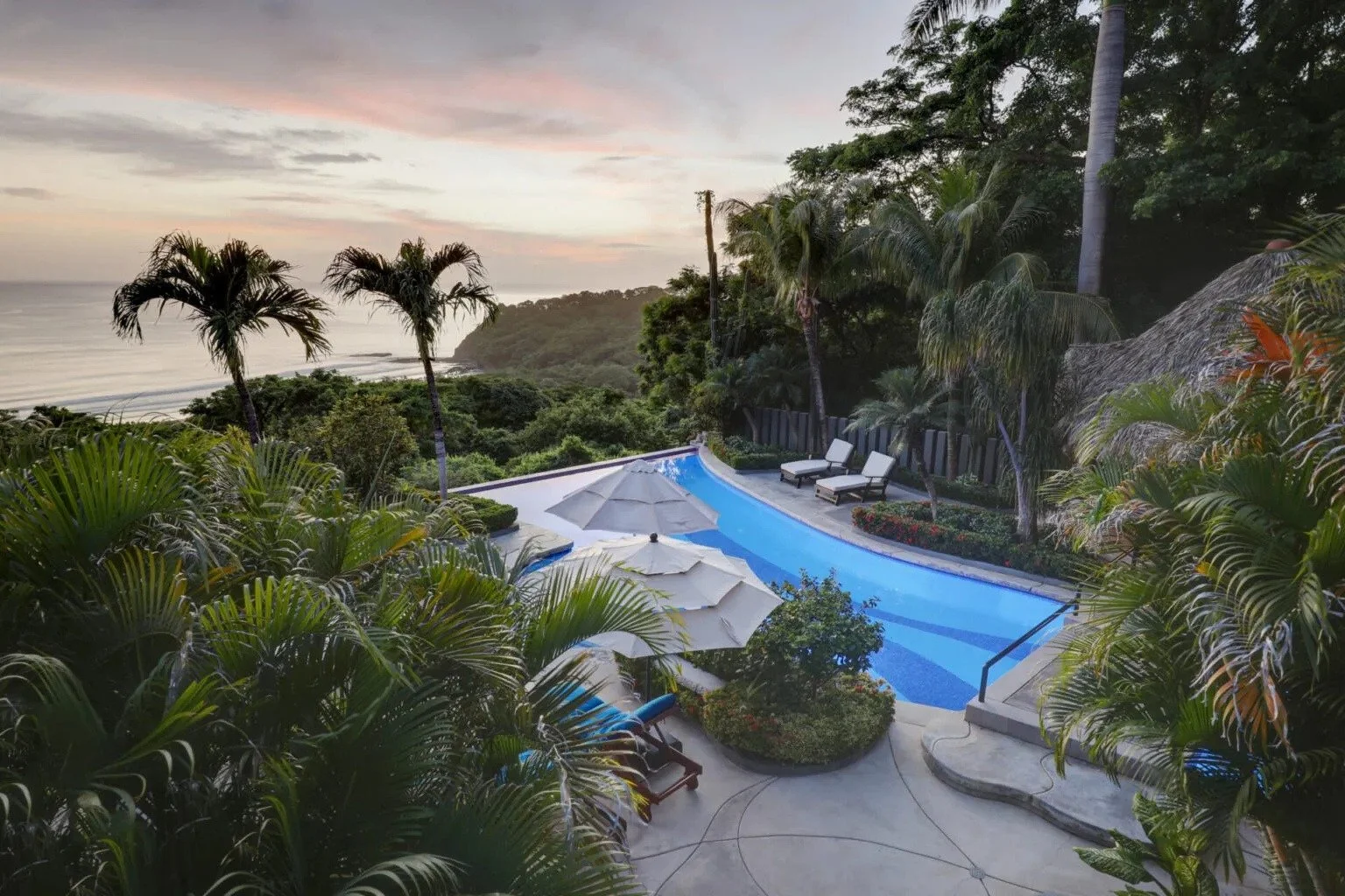 Luxury swimming pool with lounge chairs and umbrellas surrounded by lush greenery and palm trees, overlooking an ocean view at sunset.