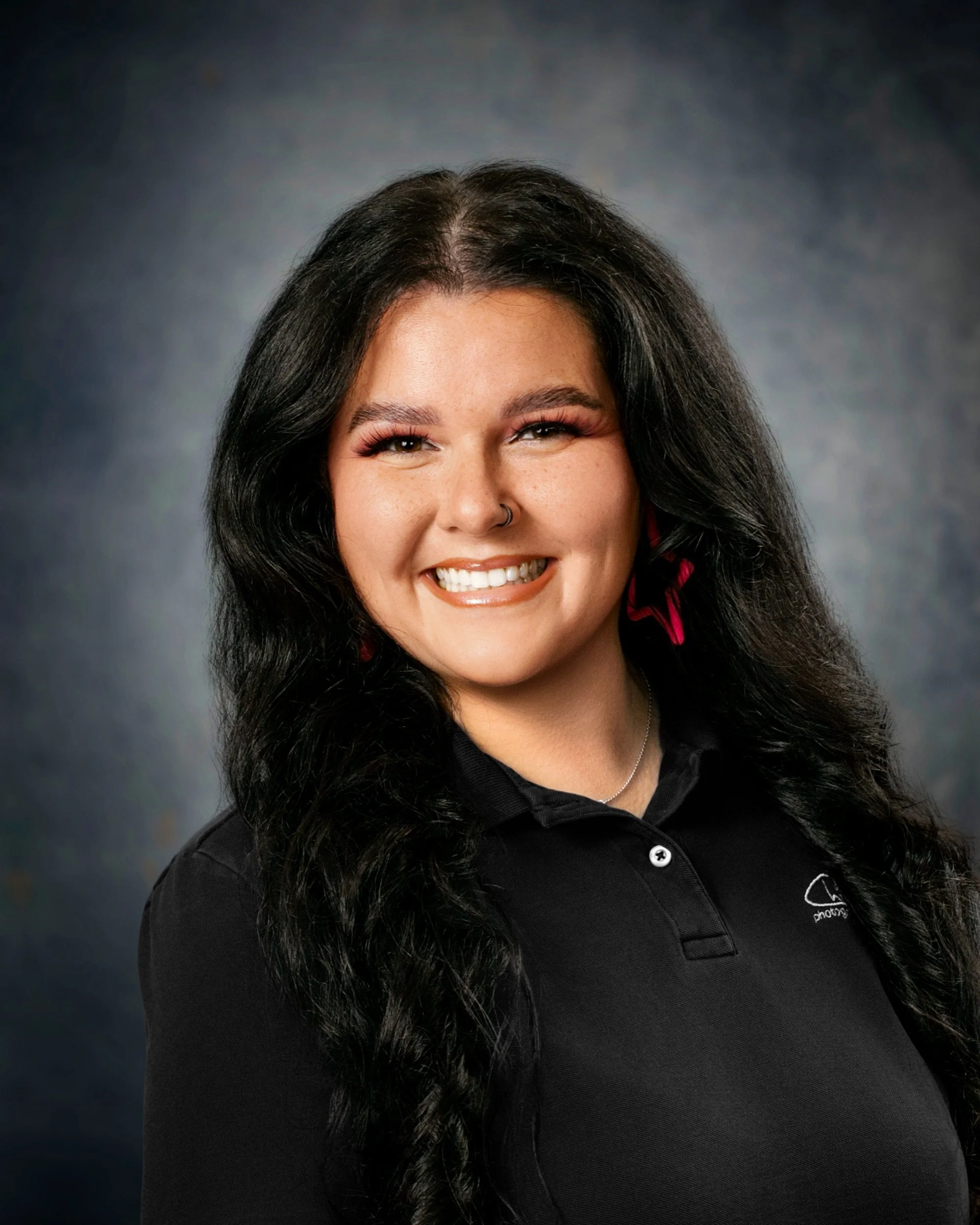 Close-up portrait of a young woman with long, dark, curly hair, smiling, wearing a black collared shirt with a logo, against a blurred, dark background.