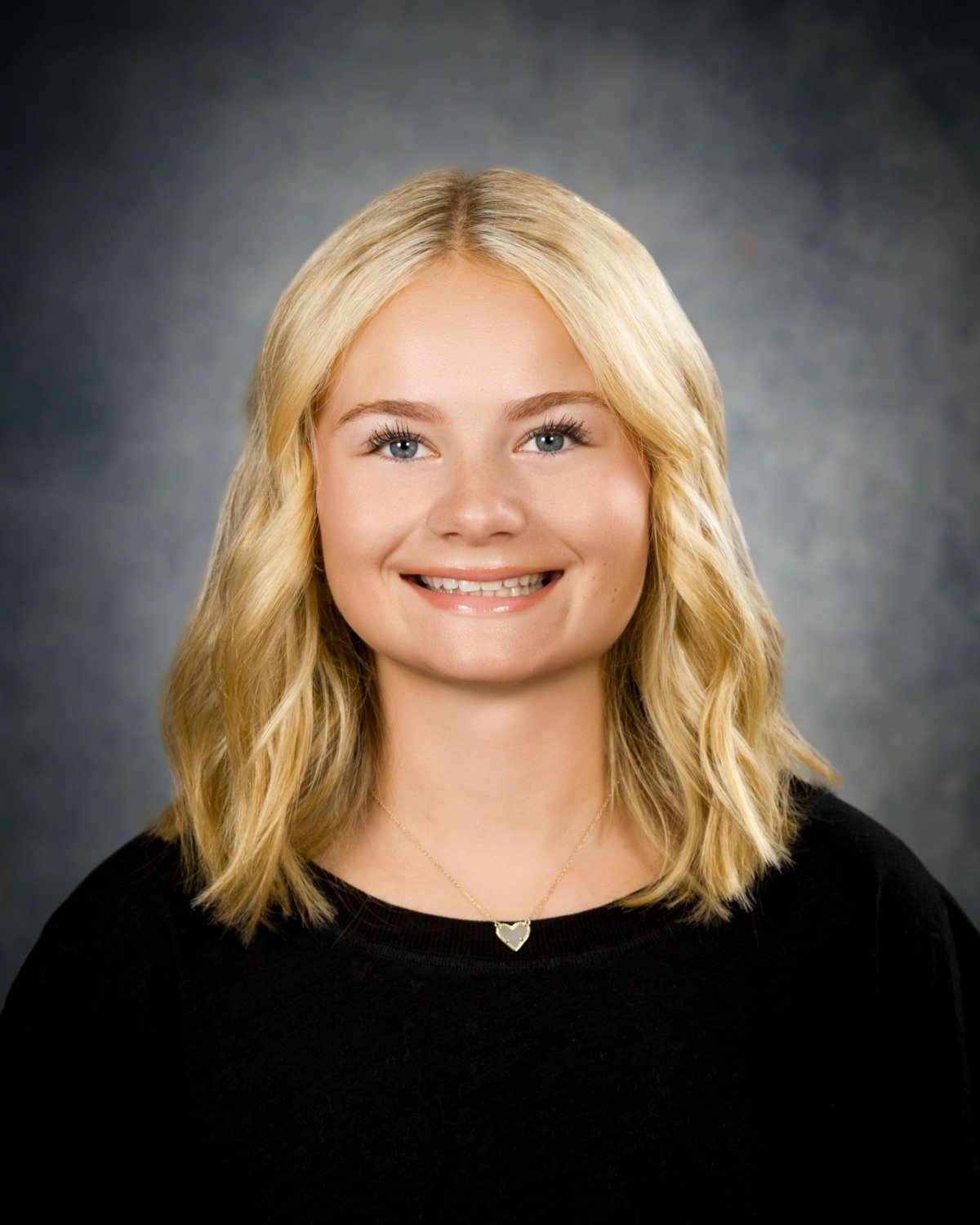 A young blonde woman with wavy hair, smiling, wearing a black top and a necklace with a heart-shaped pendant, against a gray background.