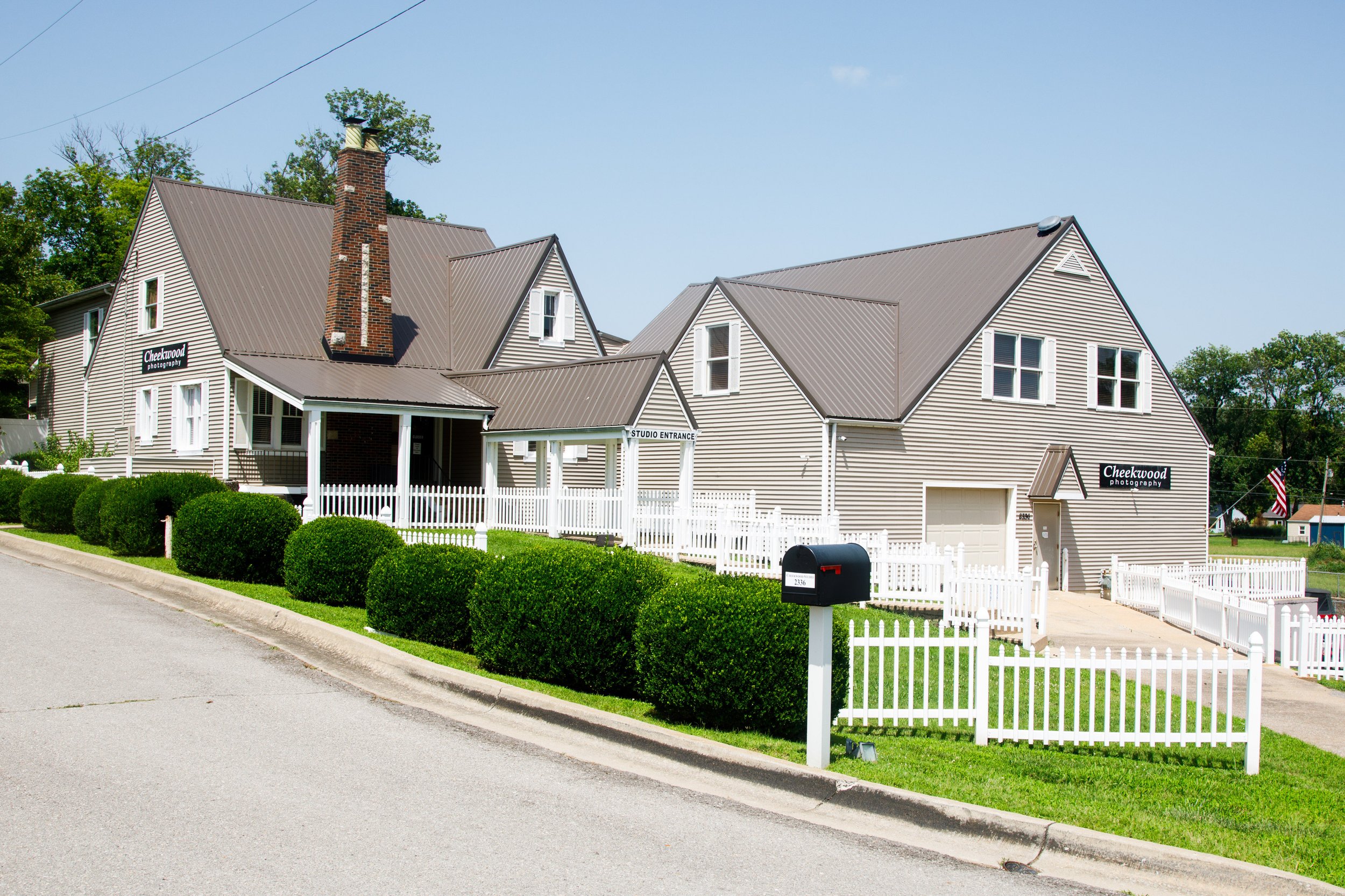 A large, two-story house with beige siding, a steep gray metal roof, and white trim. The house has multiple gables, a brick chimney, and several double-hung windows. There's a white picket fence surrounding the front yard with green bushes and a mailbox at the street. Signs indicate it is a photography studio called "Cheekwood Photography." The sky is clear and blue, with some trees in the background.
