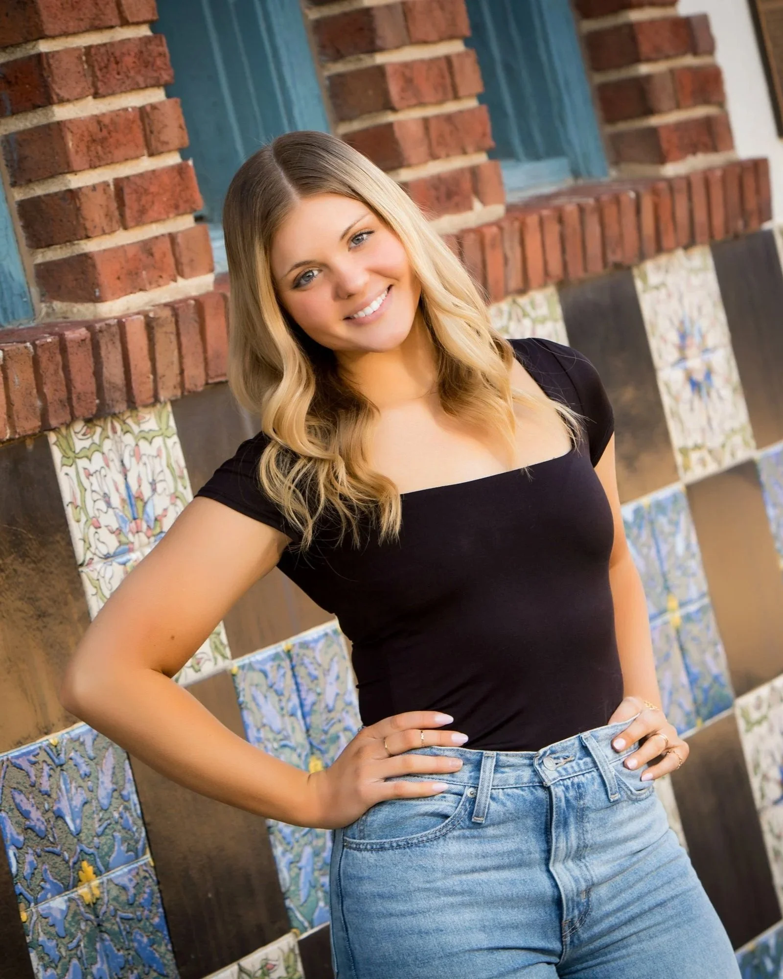 A smiling woman with blonde hair and blue eyes wearing a black top and blue jeans, standing in front of a brick and tile wall with colorful decorative tiles.