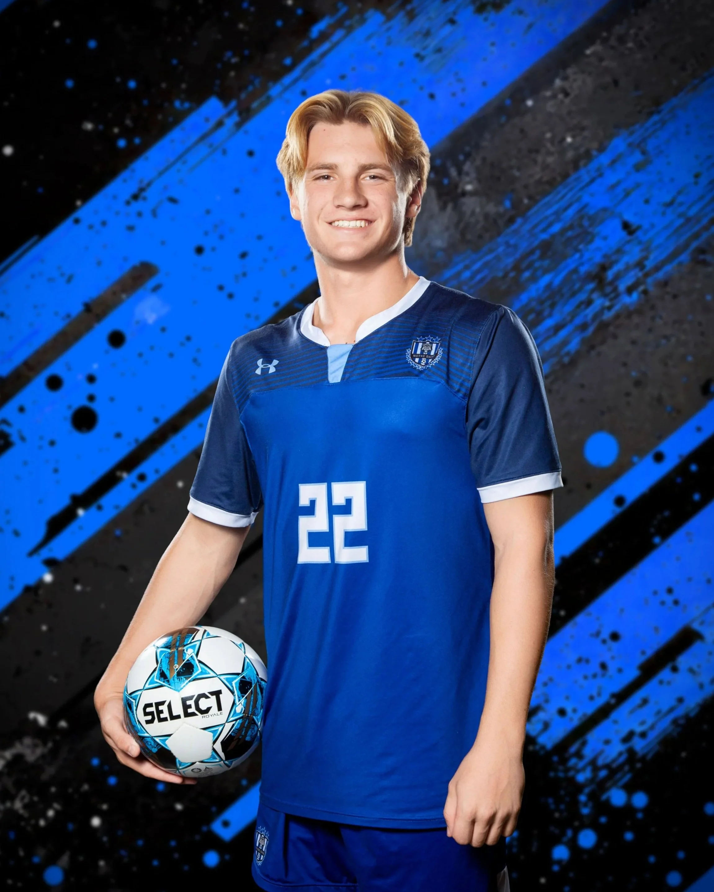 Young male soccer player in a blue uniform holding a soccer ball, smiling, with a blue and black abstract background.