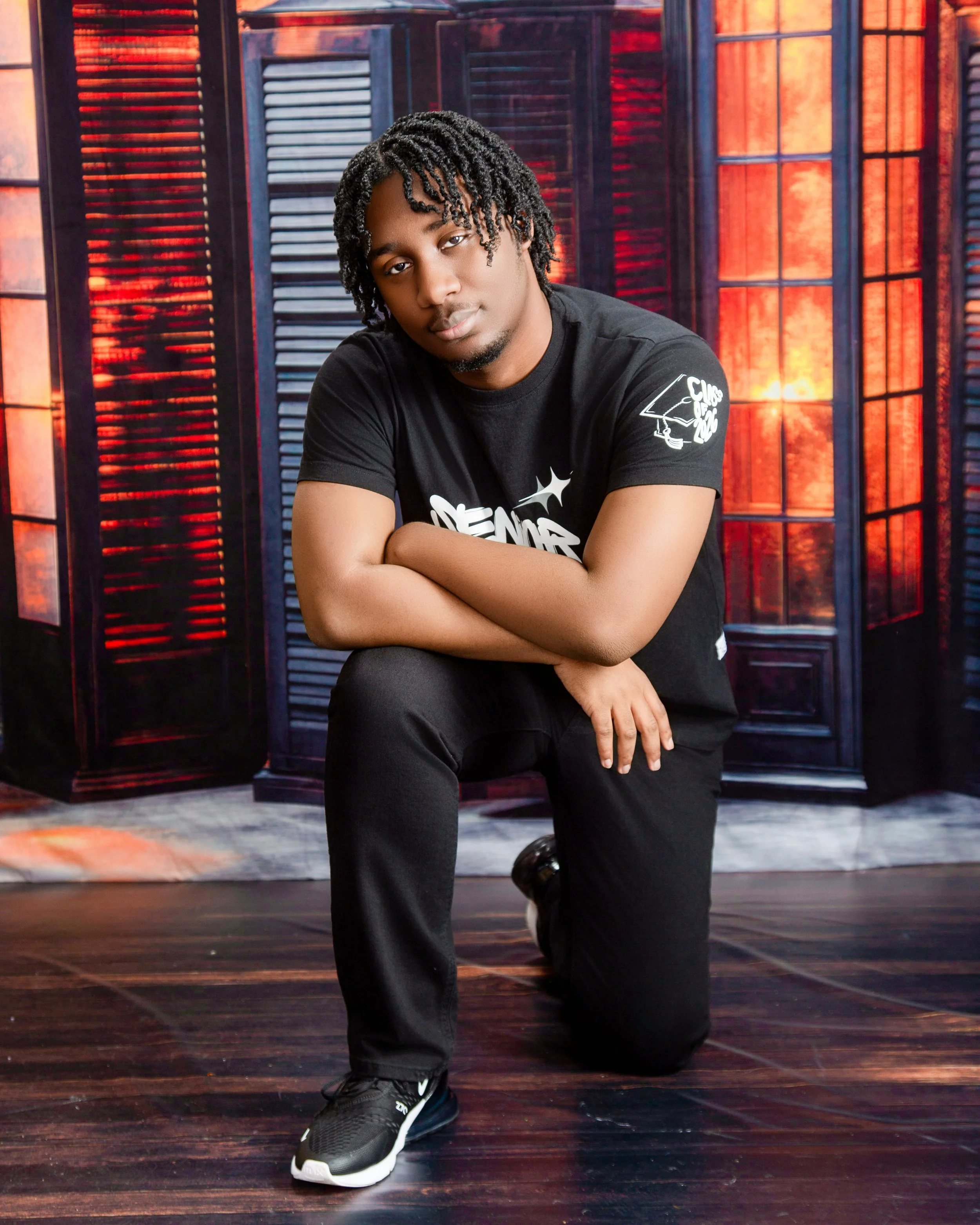 A young man with dreadlocks kneeling on one knee, wearing a black t-shirt and black pants, with a colorful wooden backdrop behind him.