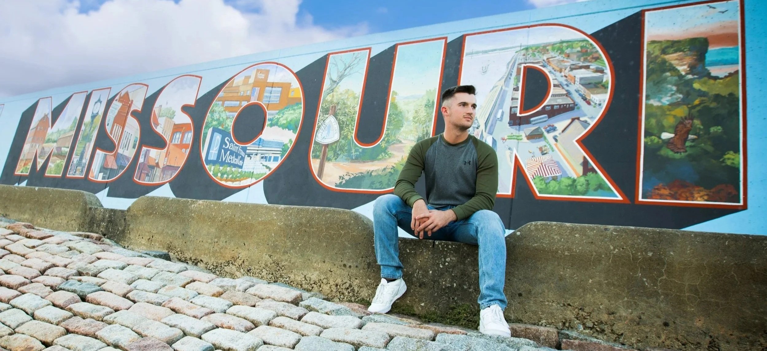 A young man sitting on a stone bench in front of a colorful mural that spells out 'MISSOURI.' The mural features scenes of a city, nature, and a river.