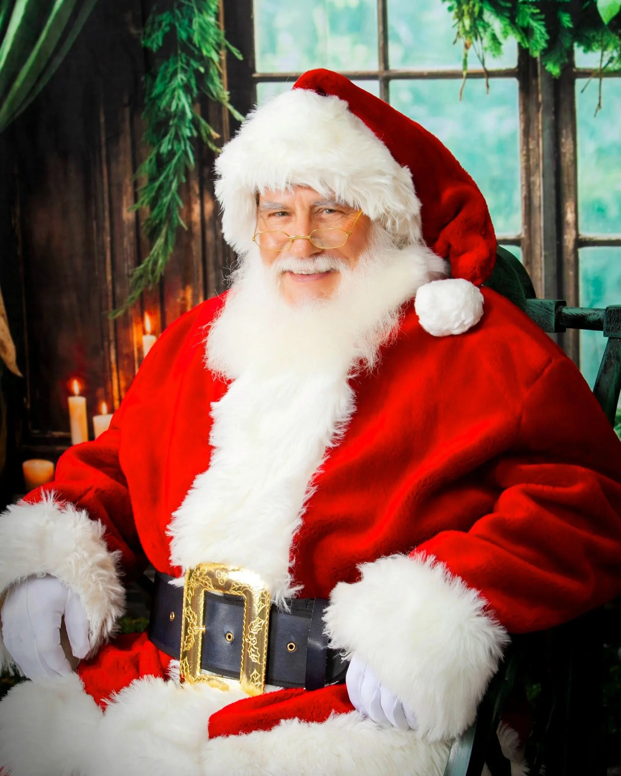 Santa Claus with glasses, white beard, red suit with white fur trim, sitting near a window with candles and greenery in the background.