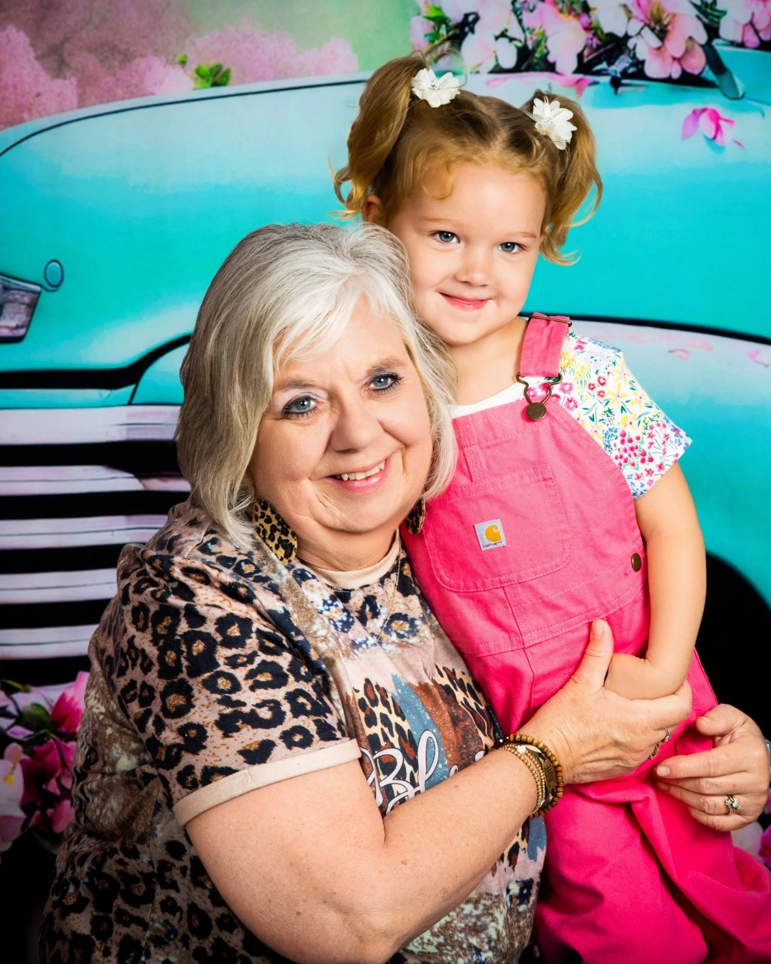 An older woman with light blonde hair and a young girl with blonde hair in pigtails and white flowers, hugging each other and smiling, with a colorful floral background.