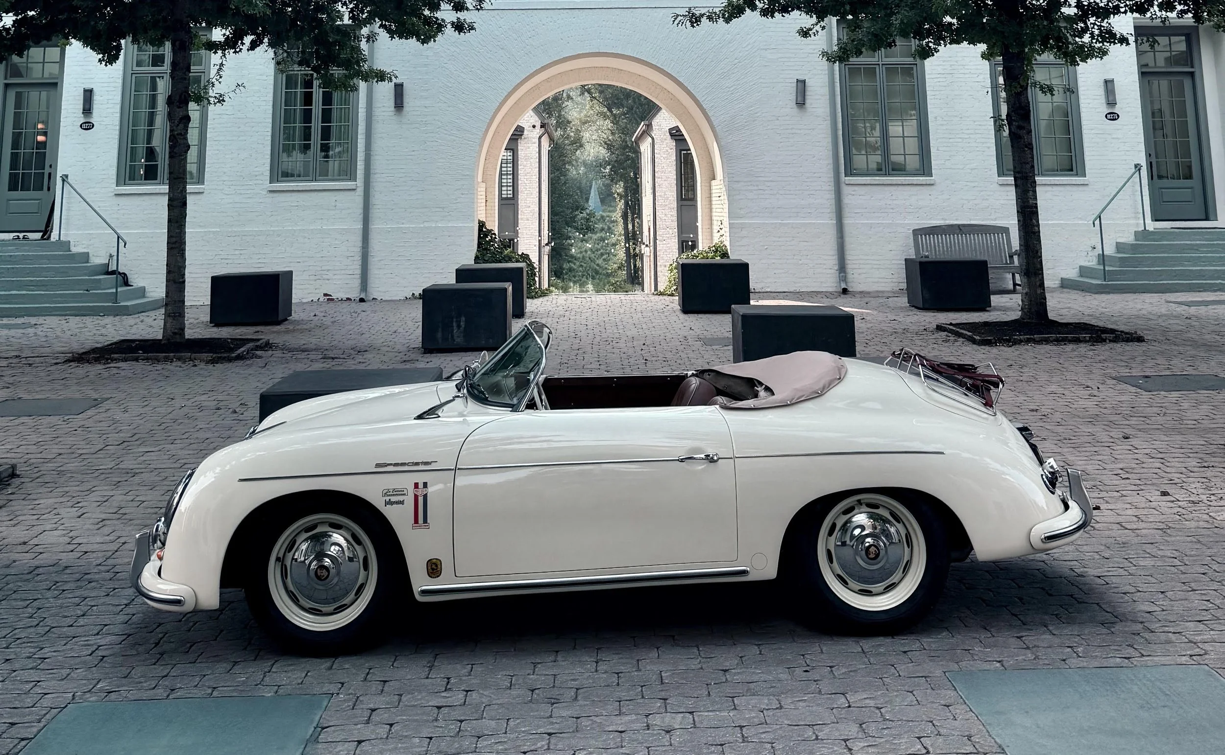 White vintage Porsche 356 Speedster owned by The Grand Exit Motorcars, parked in front of a venue with an arch and stairs, with trees and building windows in the background in Serenbe Georgia.