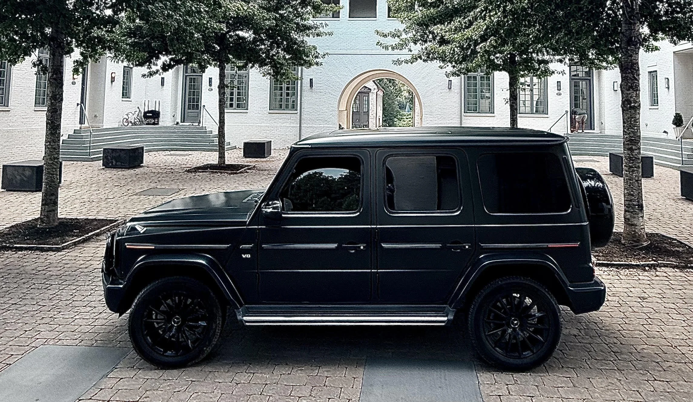 The Grand Exit Motorcars Onyx, 2020 Mercedes Benz G-Wagon 550, parked on a cobblestone street in front of a white building venue with trees and steps leading to the entrance.
