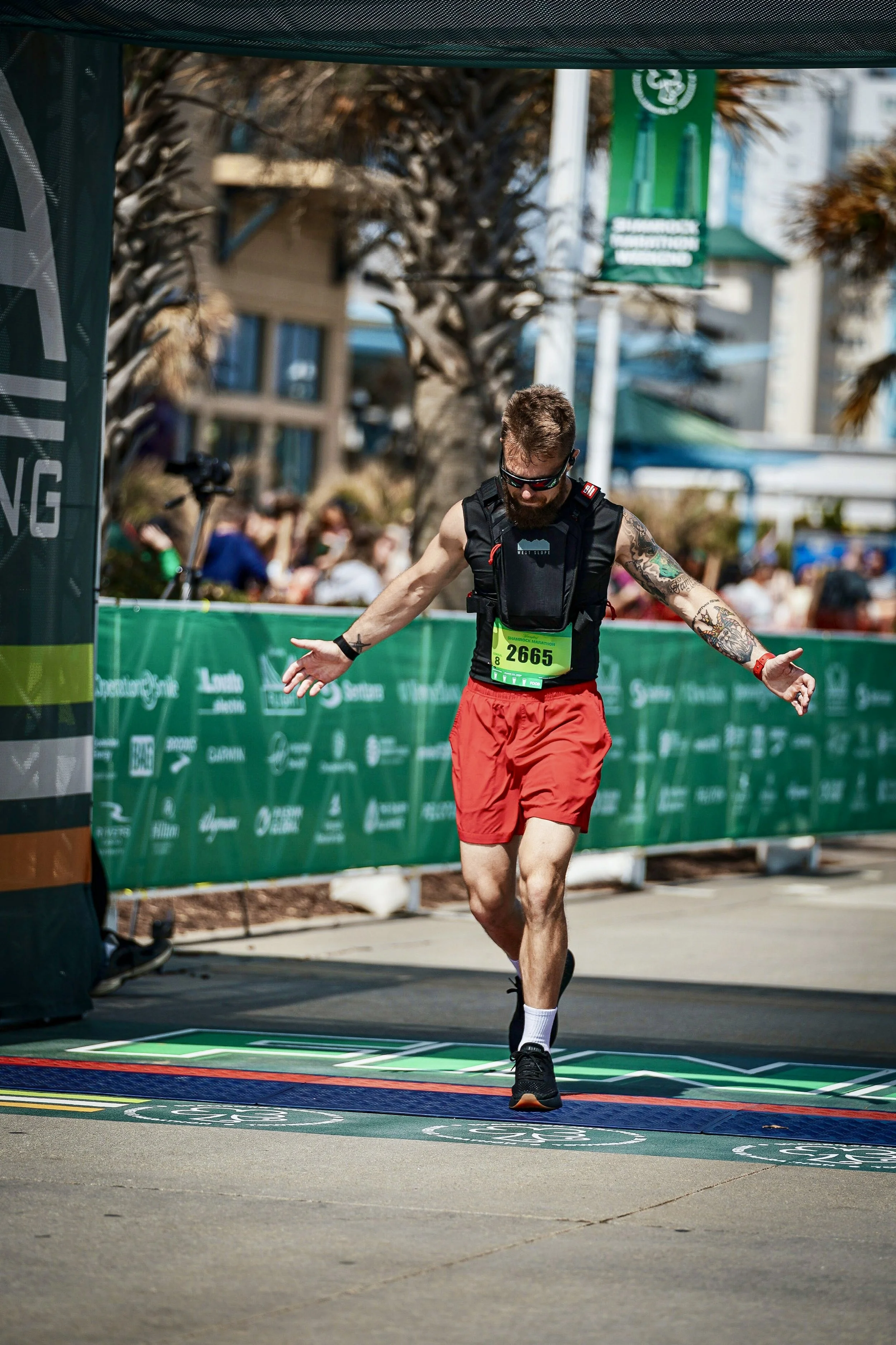 A male marathon runner crossing the finish line with arms outstretched, wearing black sunglasses, a black vest, red shorts, and black running shoes. He has visible tattoos on his arms and a green race bib numbered 2665. There are spectators and trees in the background, along with banners and a green finish line mat.