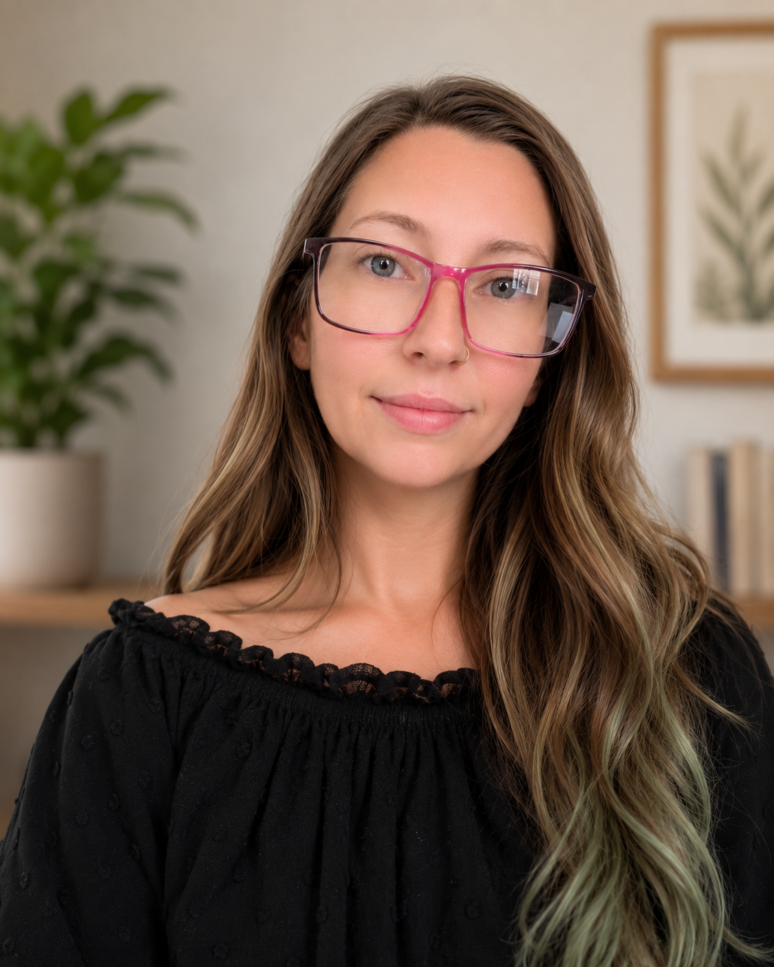 A woman with long wavy hair, wearing pink glasses and a black off-shoulder top, standing indoors with a blurred background of a plant and framed art.