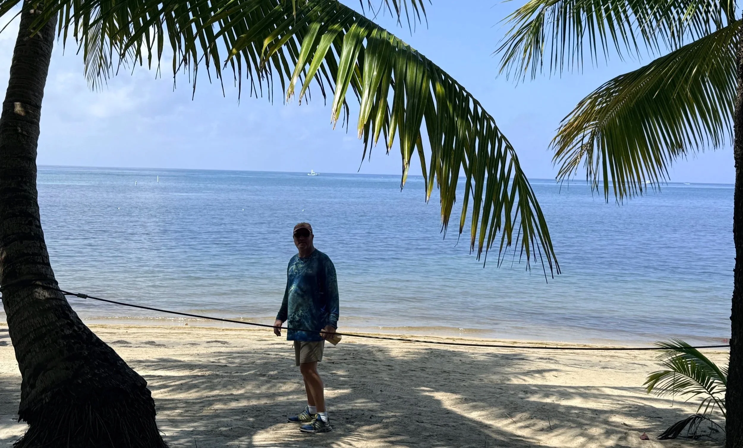 Man standing under a palm tree on a beach with the water behind him