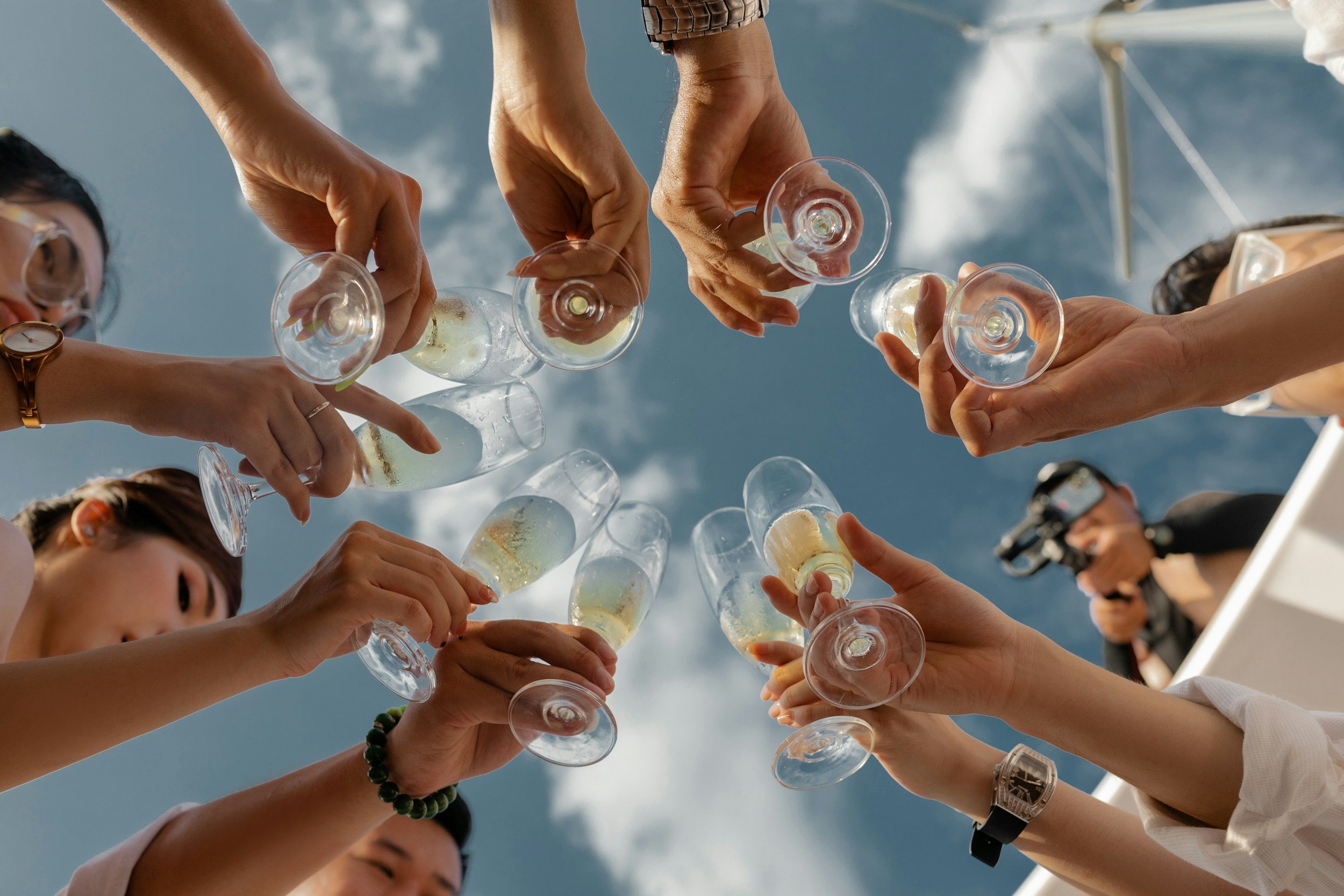 Multiple people are raising glasses filled with champagne for a toast outdoors against a blue sky with clouds. Some individuals are wearing watches, bracelets, and sunglasses.