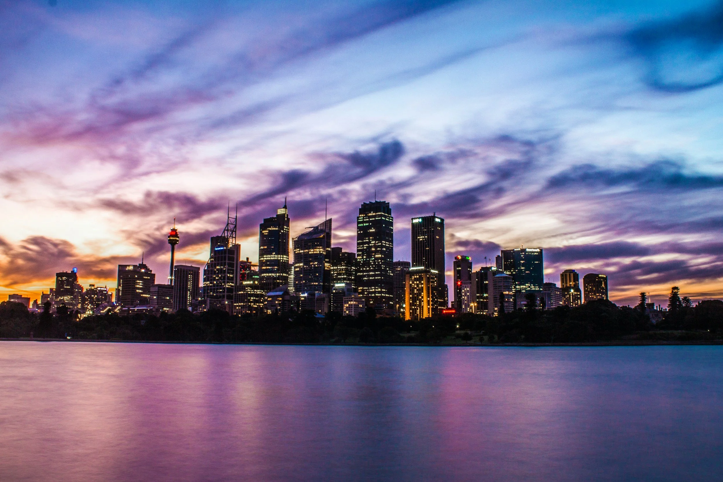 A city skyline at dusk with tall buildings and skyscrapers, a water body in the foreground, and colorful clouds in the sky.