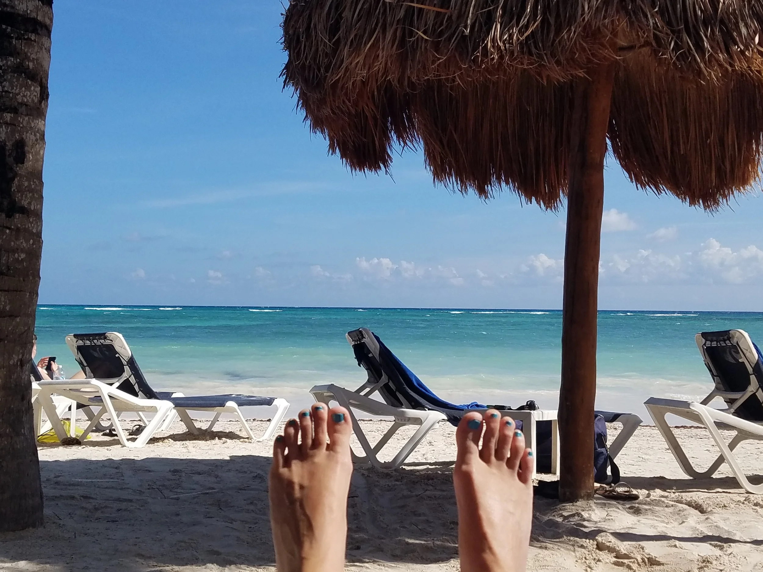 A sandy beach with lounge chairs, palm trees, and a straw umbrella. The ocean and a blue sky with some clouds are visible in the background. A person's legs with painted toenails are visible in the foreground.