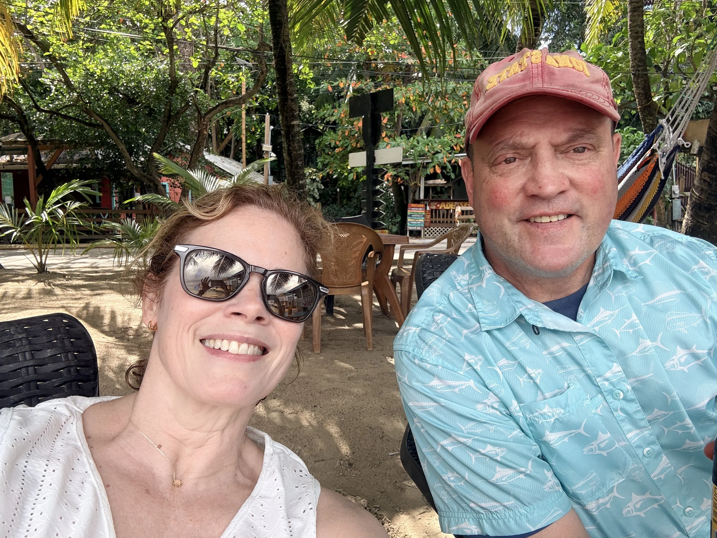 A smiling woman with sunglasses and a man in a pink cap, both partially visible, sitting outdoors under trees on a sunny day at a beachside or tropical location.