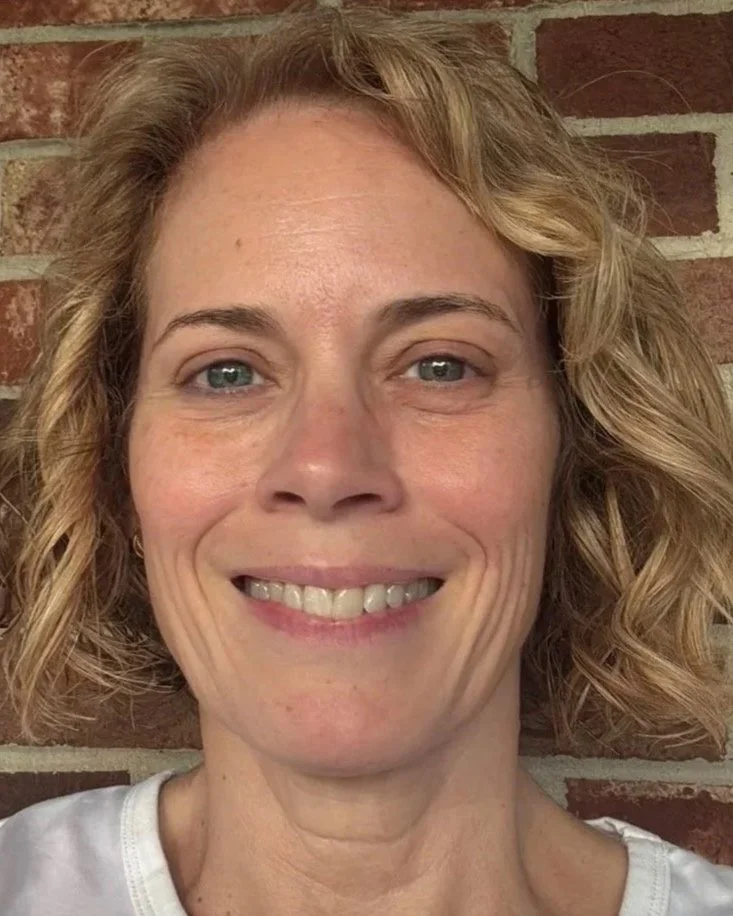 Close-up of a smiling woman with short, curly blonde hair, blue eyes, and light skin, standing against a brick wall background.