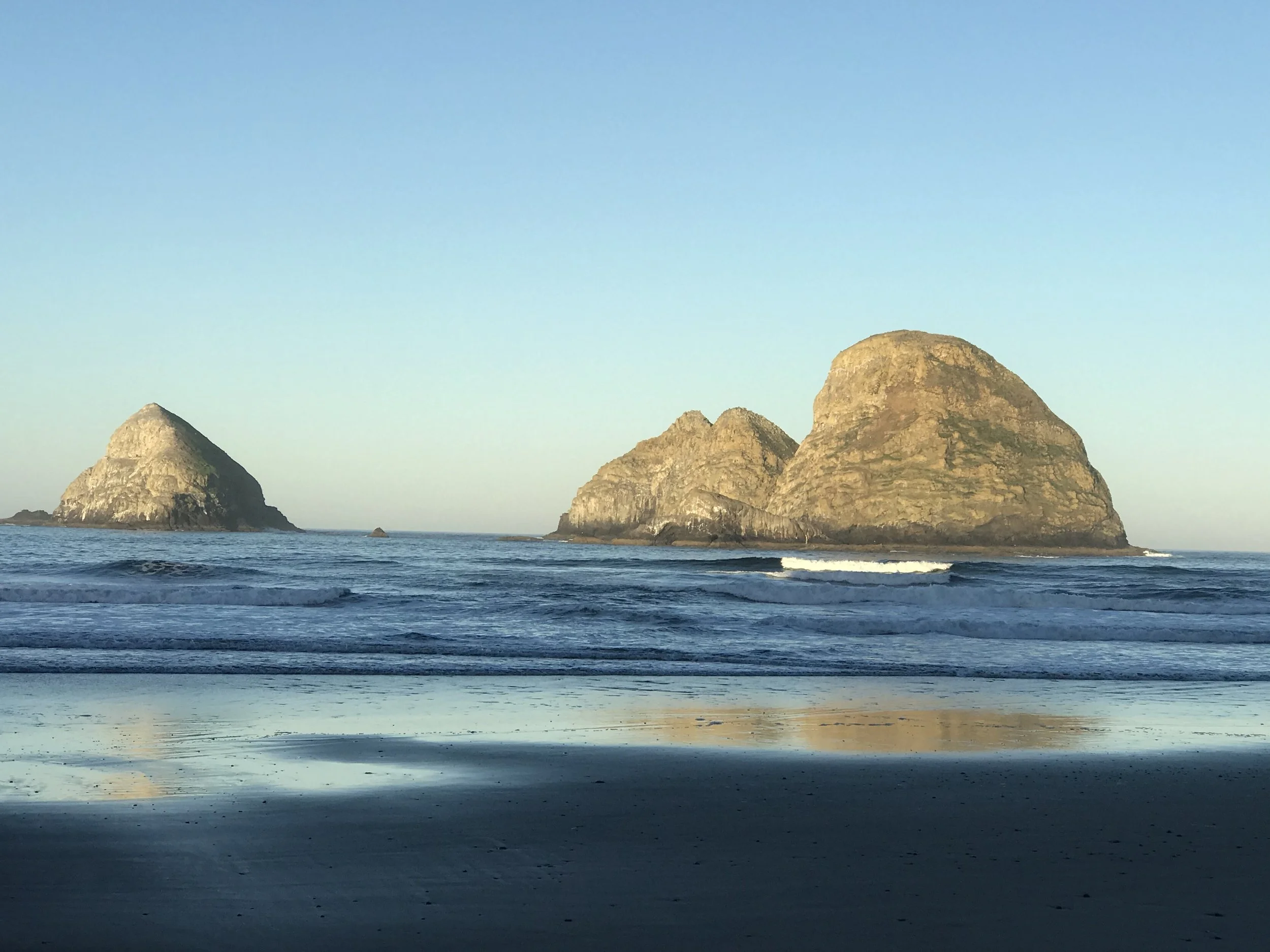 A seaside view with large rocky islands in the distance under a clear blue sky, calm ocean water, and a sandy beach in the foreground.