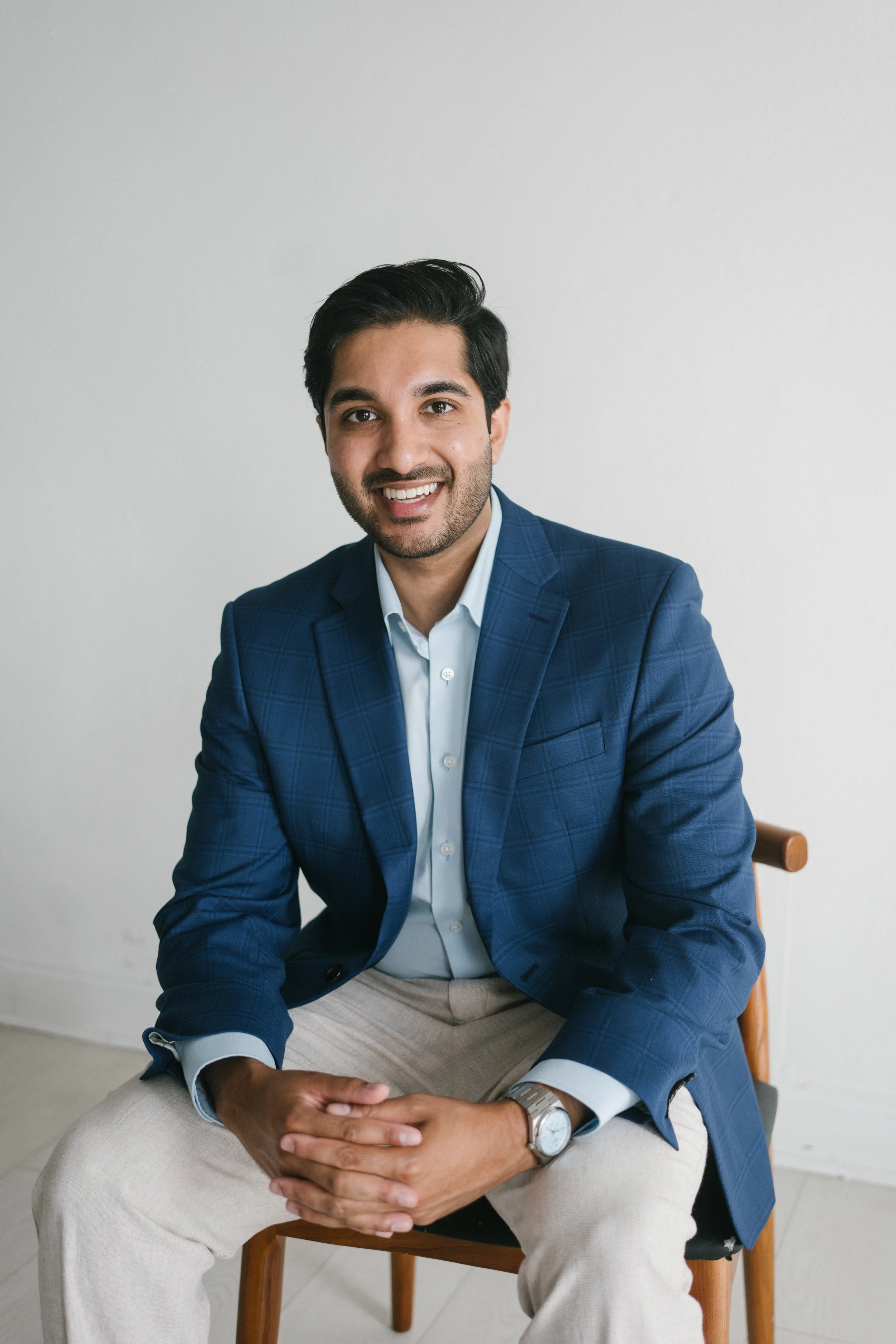 A man with dark hair, beard, and mustache wearing a blue checked blazer, white shirt, and beige pants sitting on a wooden chair against a plain white wall.