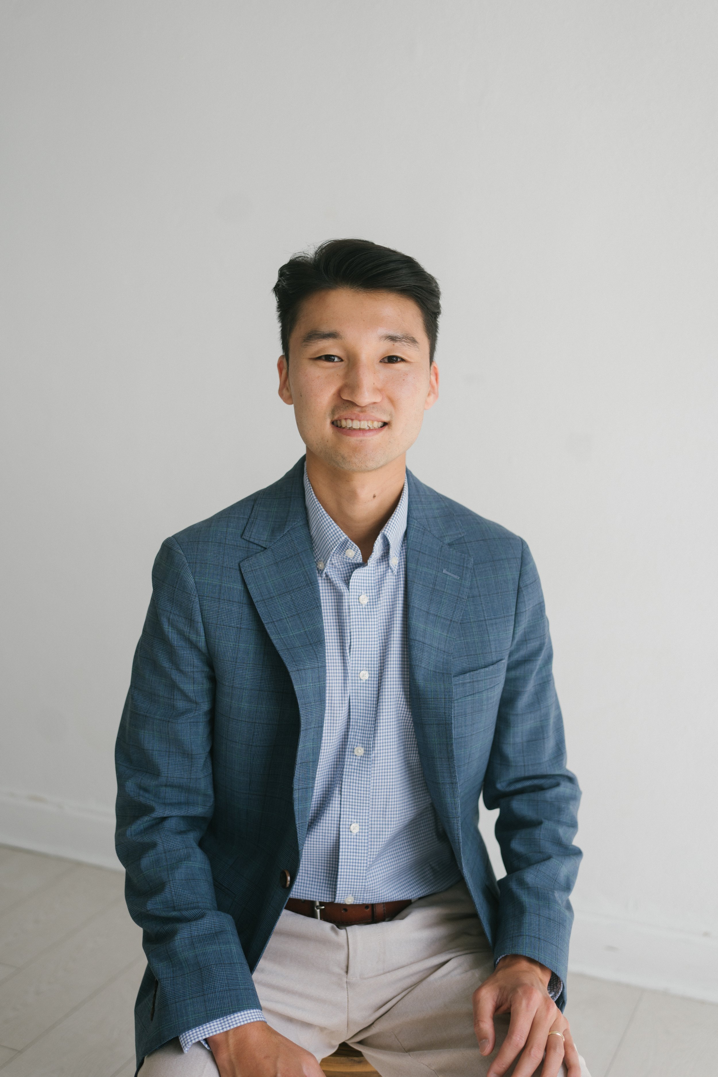 Portrait of a young man wearing a blue plaid blazer, light blue shirt, and beige pants, sitting on a wooden stool against a plain white wall.