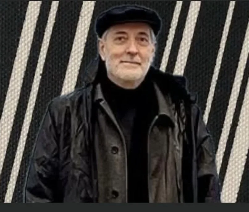 An older man with a gray beard wearing a black cap, black jacket, and black shirt, standing in front of a geometric-patterned black and white background.