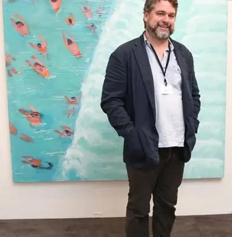 A man with a beard and glasses stands in front of a painting depicting swimmers in the water.