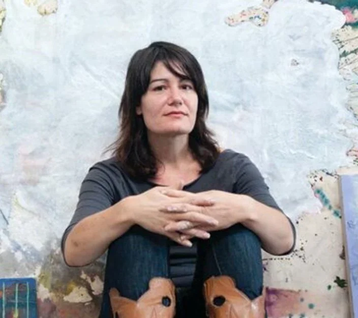 A woman with shoulder-length dark hair sitting against a graffiti-covered wall, wearing a gray top, blue jeans, and brown boots.