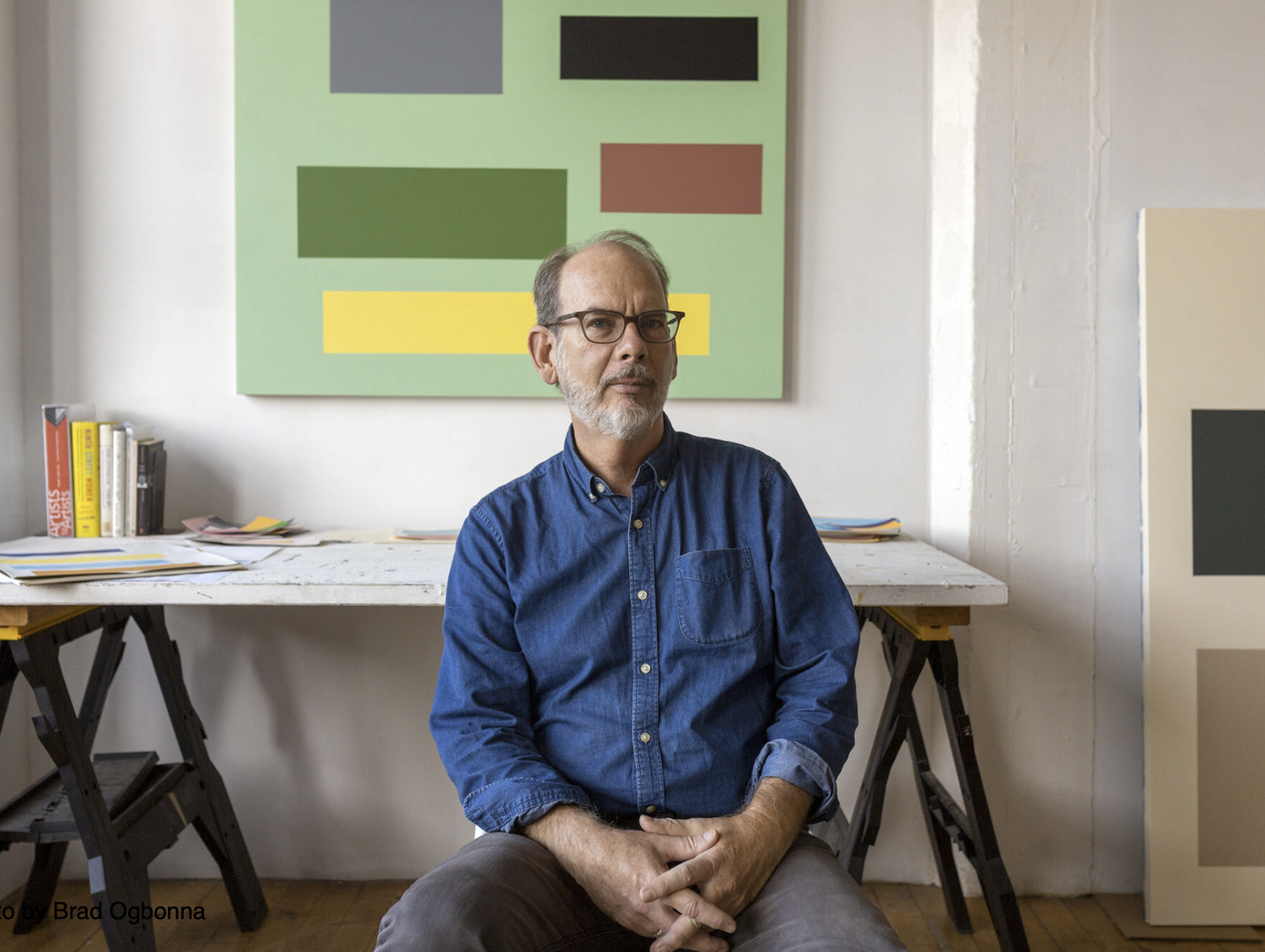 Middle-aged man with glasses and a beard, wearing a blue button-up shirt, sitting on a stool in an art studio. Behind him, abstract artwork with blocks of green, gray, black, red, and yellow on a white wall, and a table with books and papers.