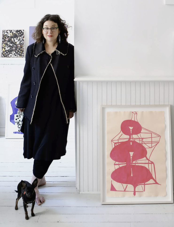 A woman with curly dark hair wearing glasses, a black coat, and a black dress, standing inside an art gallery next to a small black dog. There are colorful abstract artworks on the white wall behind her, and a large framed pink artwork with geometric shapes leaning against a white paneled wall.