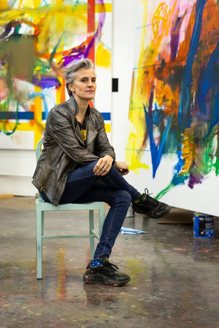 A woman with short gray hair sits on a chair in front of an abstract painting in an art studio, wearing a metallic jacket, dark jeans, and black sneakers, with her foot resting on another chair.