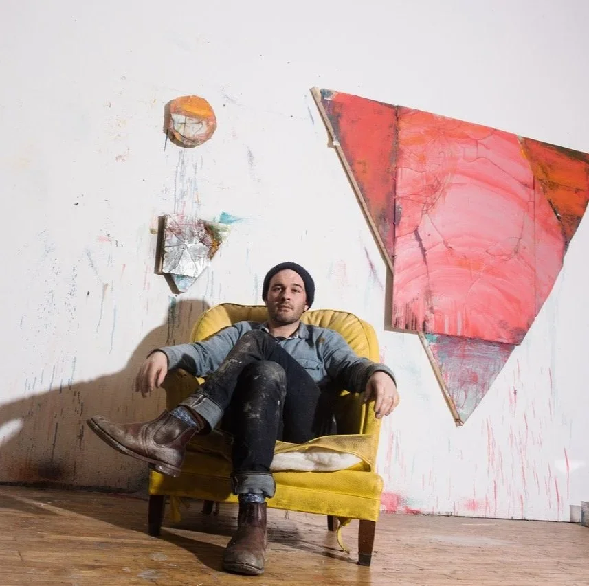 A man sitting on a yellow armchair in a room with an abstract painting on the wall. The wall has a colorful, irregular pattern with splashes of paint.