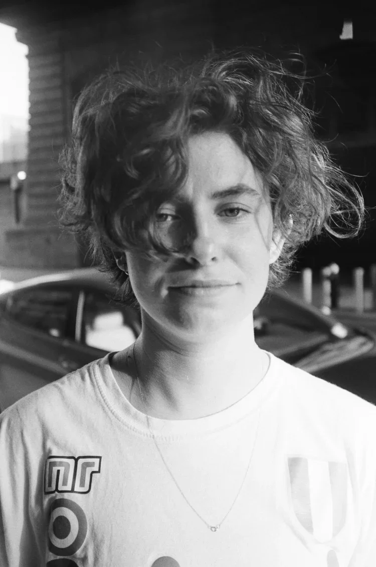 A young person with curly, tousled hair smiling slightly for a black-and-white photo, wearing a T-shirt with graphics and a necklace, standing outdoors near a car and a building.