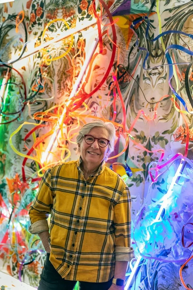 Older woman with glasses and grey hair smiling, standing in front of a colorful, neon art installation with swirling ribbons of light and floral patterns.