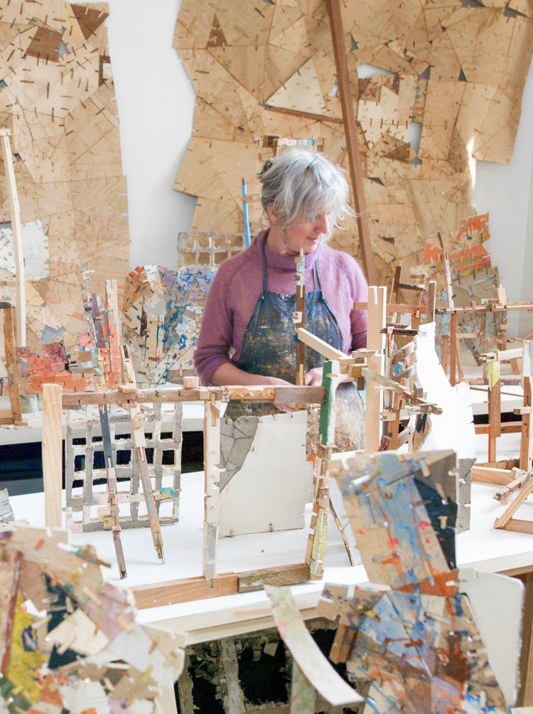 An elderly woman with gray hair wearing a purple sweater and an apron working on a wooden art piece in a studio filled with various abstract wooden artworks and projects.