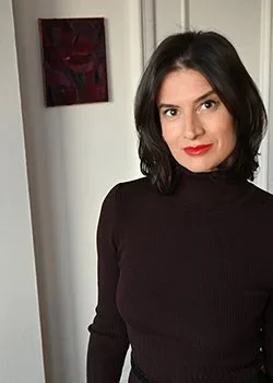 A woman with shoulder-length black hair and red lipstick wearing a black turtleneck sweater, standing indoors near a white door with a red and black abstract painting on the wall behind her.