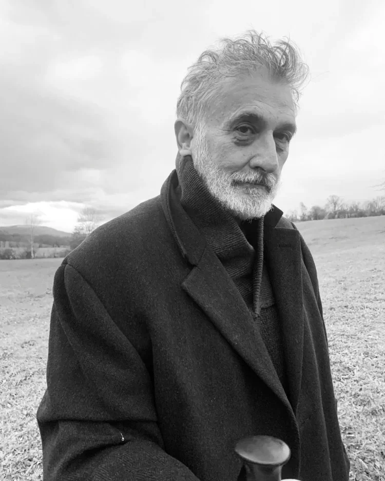 A black and white photo of an older man with a beard and curly hair, wearing a dark coat, standing outdoors in a grassy field with cloudy sky in the background.