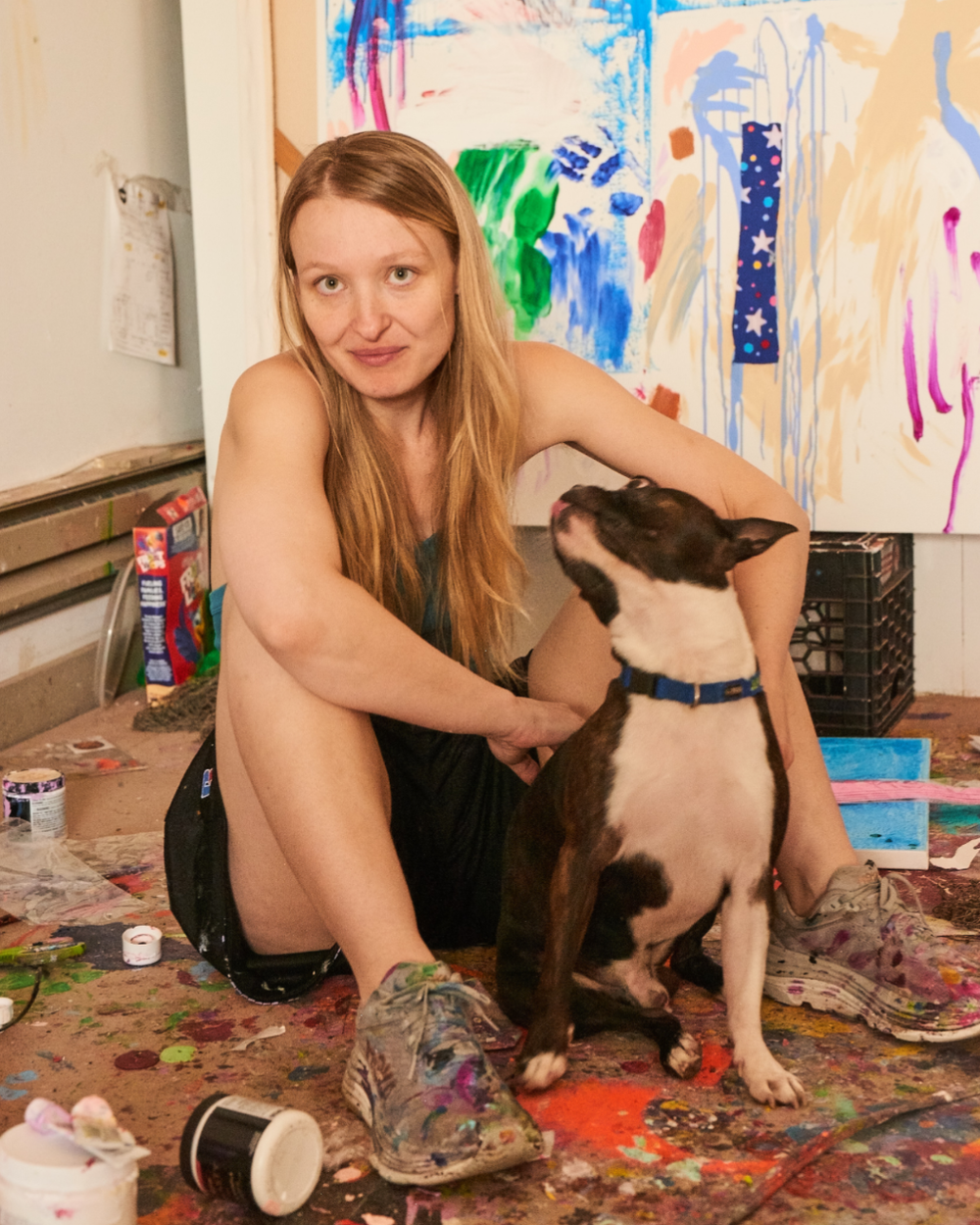 A woman with long red hair sitting on the floor with a Boston Terrier dog in an art studio with colorful, expressive paintings on the walls. The floor is covered with paint splatters and art supplies.