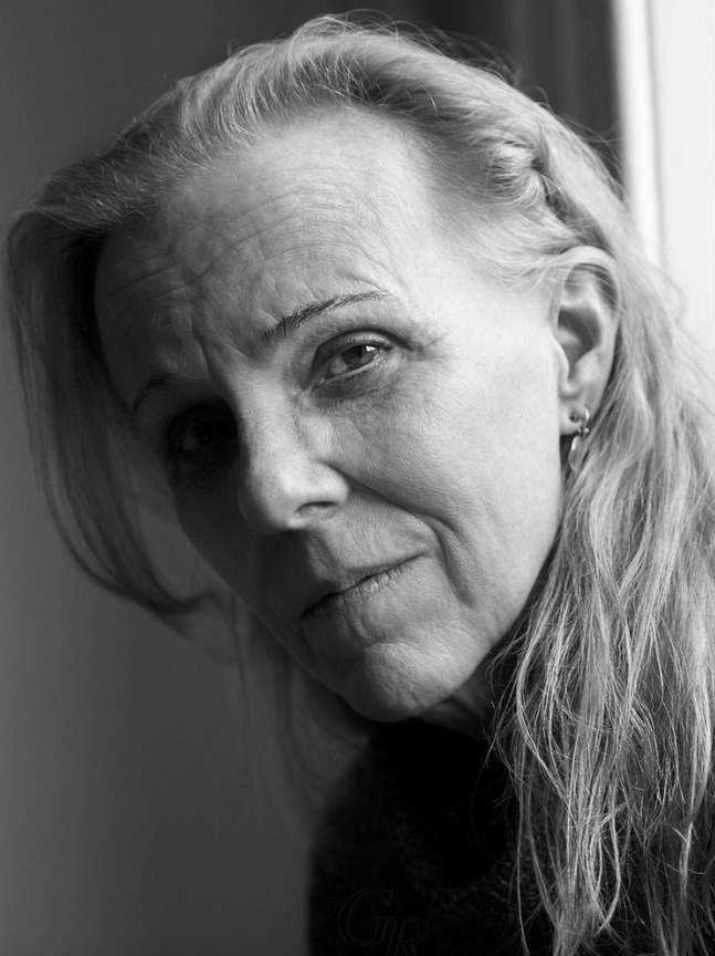 Black and white portrait of a mature woman with long, light hair, gazing thoughtfully at the camera near a window.