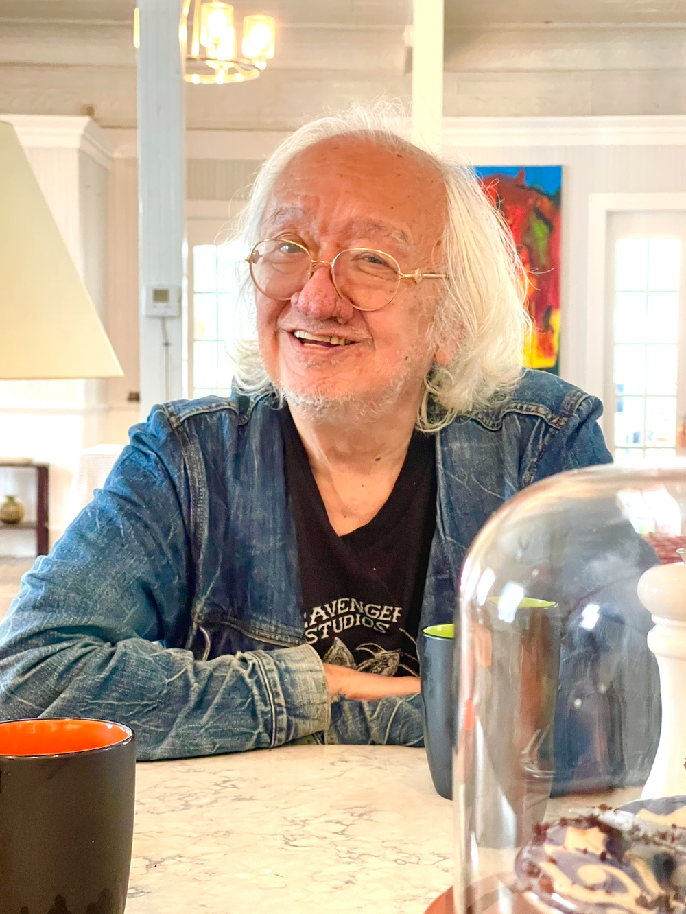 An elderly man with white hair, glasses, and a beard, smiling at a table with cups, a cake, and a glass jar in a cozy, well-lit room with large windows and colorful artwork on the wall.