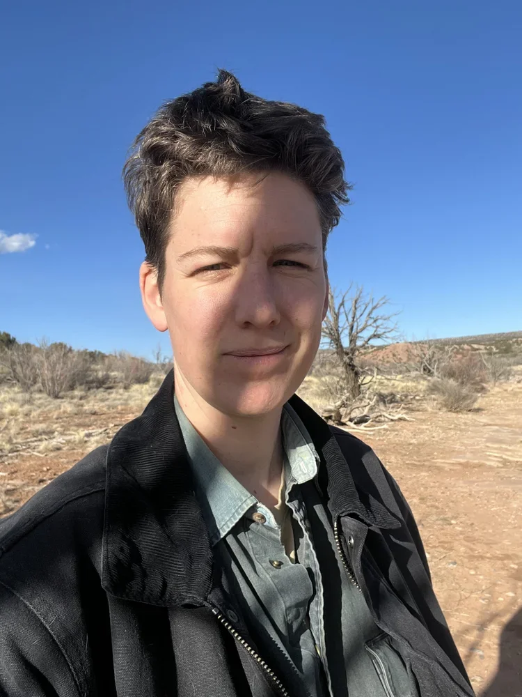 A person standing outdoors on a sunny day with a clear blue sky, dry landscape, and leafless trees in the background.