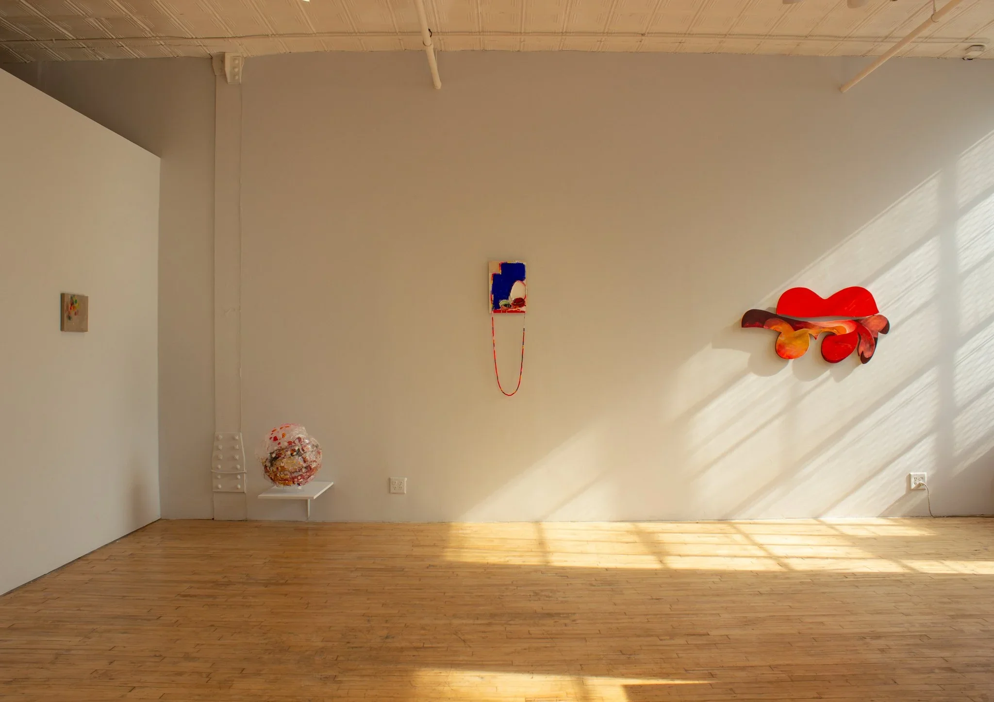 Empty art gallery with white walls, decorated with colorful wall art, sculptures, and a small white stand. Natural light creates shadows on the wooden floor.