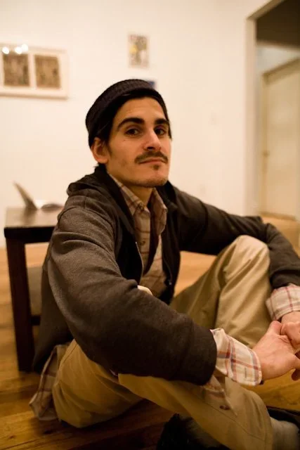 A man sitting on the floor inside a room, looking at the camera with a serious expression, wearing a black hat, a dark jacket, and beige pants.