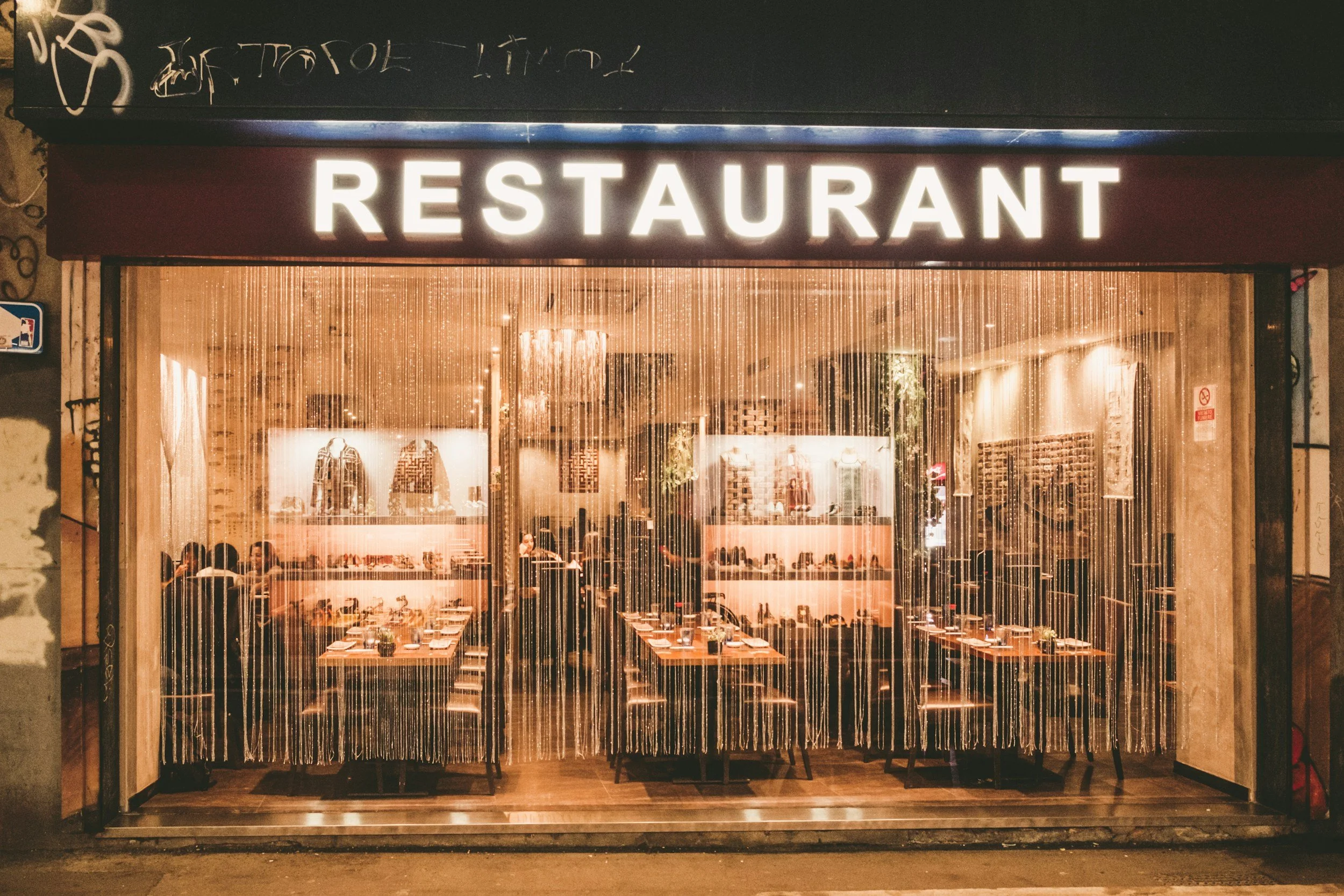 Restaurant storefront with large illuminated sign reading 'RESTAURANT' above a glass window showing the interior dining area at night.