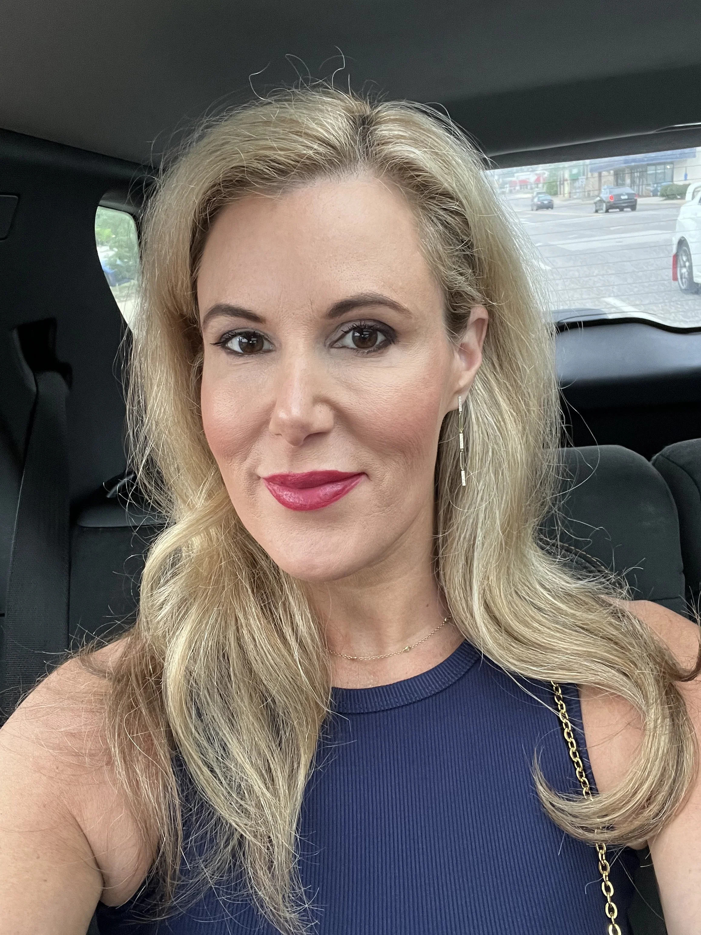 A woman with blonde hair, makeup, and red lipstick posing for a selfie inside a vehicle, wearing a blue sleeveless top and a gold chain strap purse.