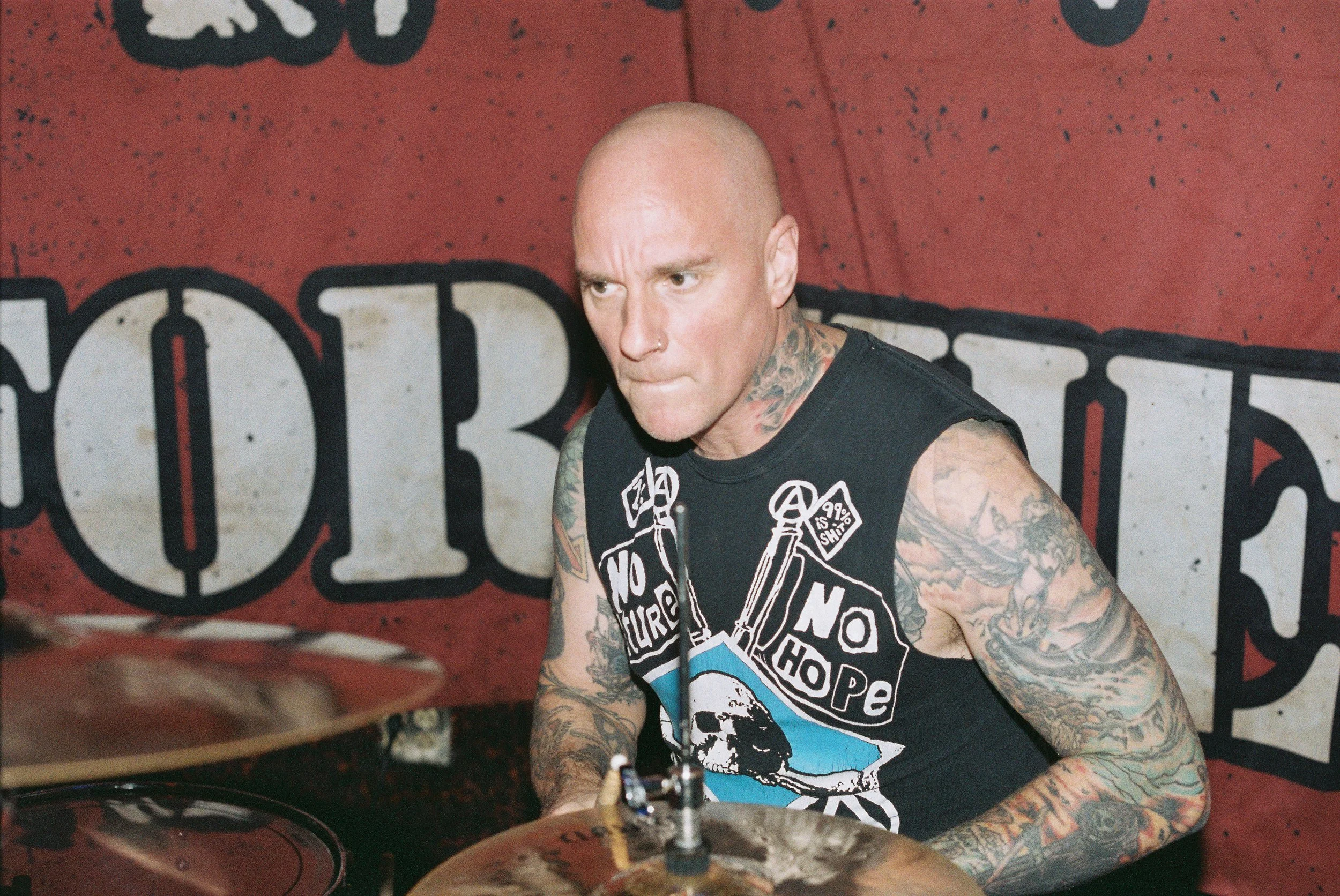 A tattooed man with a shaved head wearing a sleeveless black T-shirt, sitting at a drum set in front of a red wall with black and white graffiti.