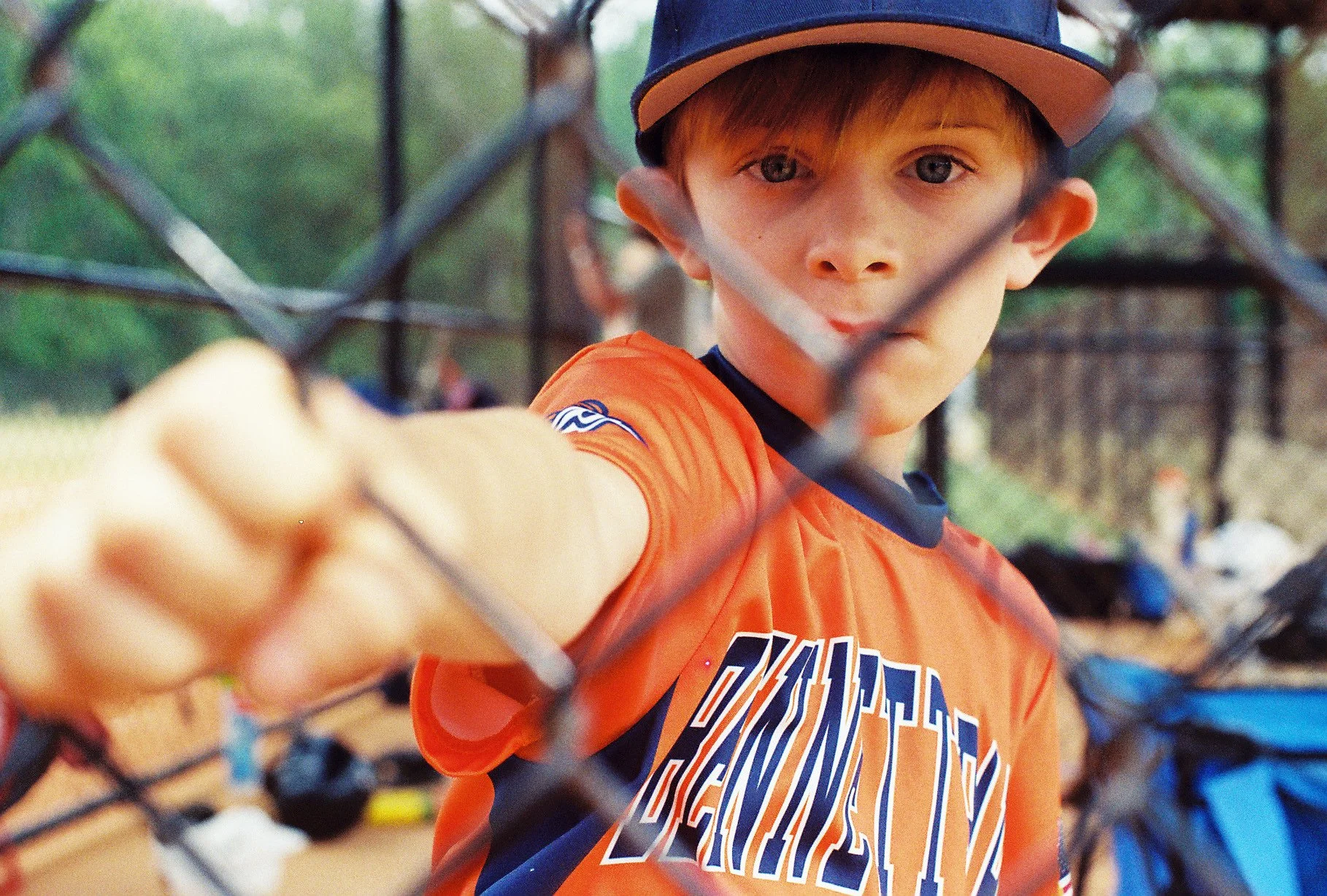 A young boy with light brown hair and blue eyes wearing an orange T-shirt and a blue and orange baseball cap, standing behind a chain-link fence and looking through it with his finger extended towards the camera.
