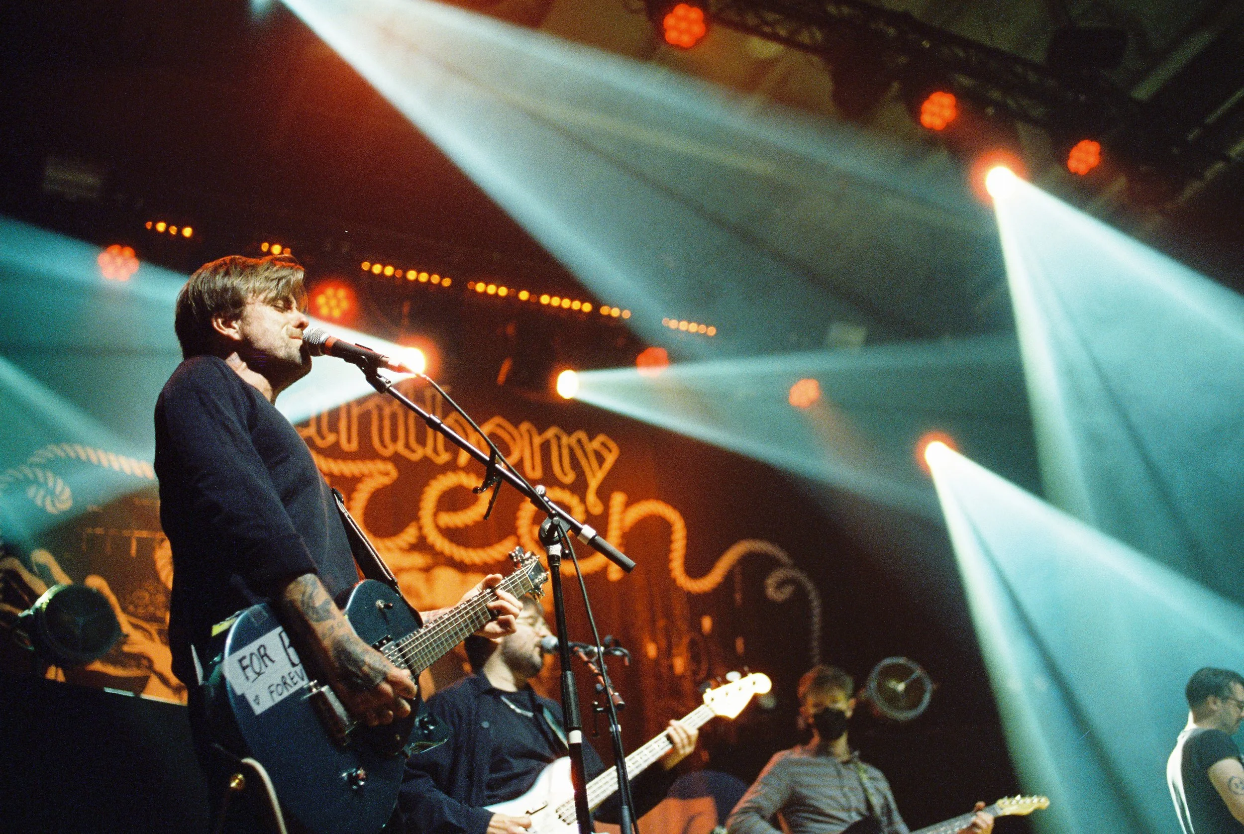 A musician with tattoos on his arm performing on stage with a microphone and guitar, while other band members play guitars in the background under stage lights.
