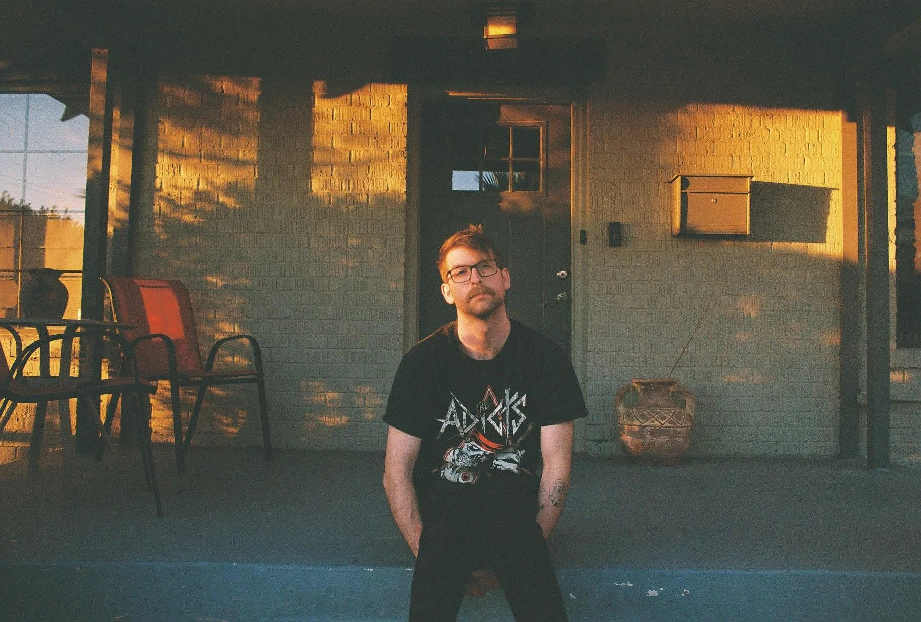 A man with glasses and tattoos is sitting on a porch in front of a brick wall. The porch has several chairs and a potted plant, with warm sunlight casting shadows on the wall.