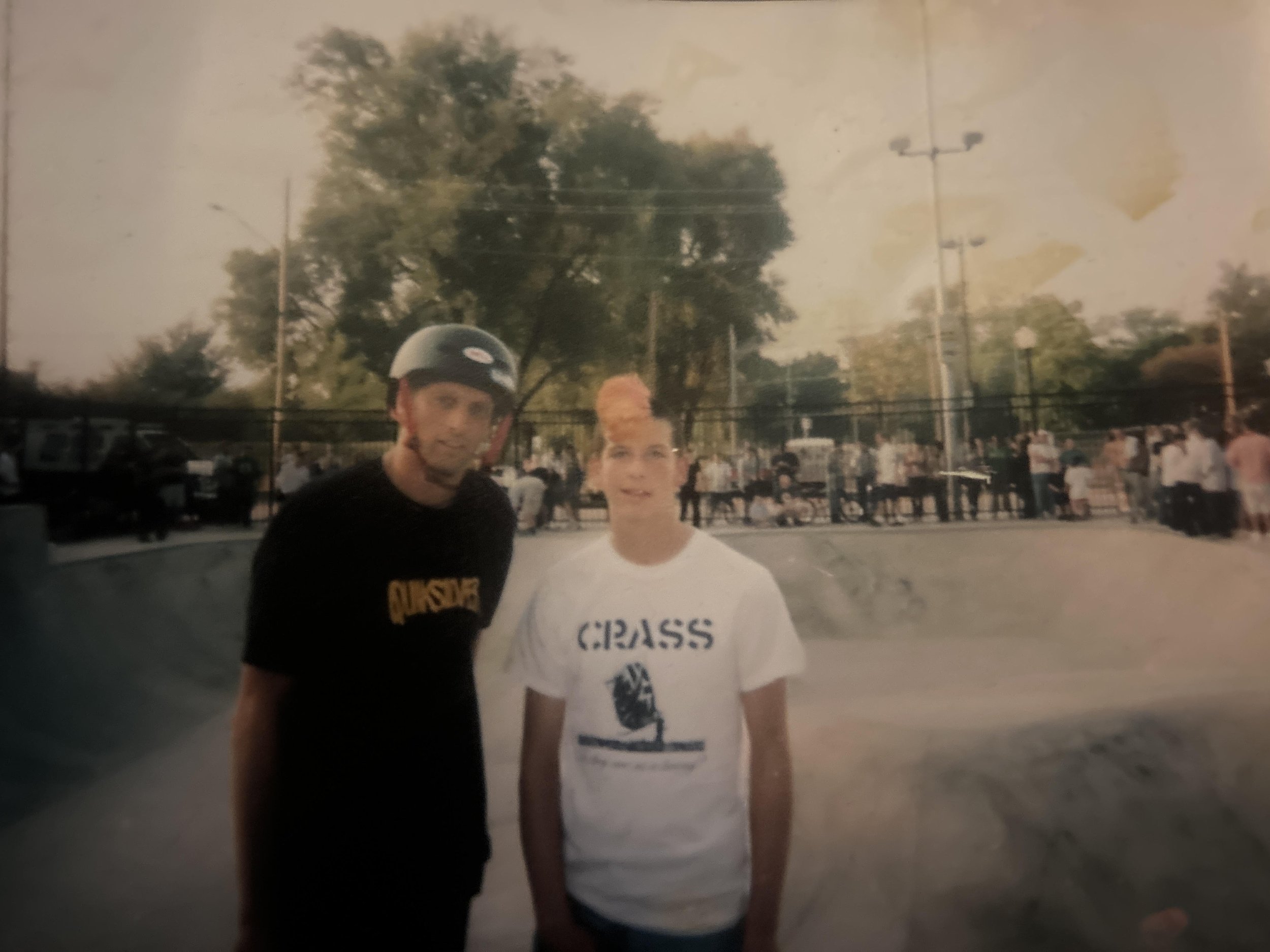Two young men standing outdoors during daytime, one wearing a helmet and the other wearing a white t-shirt that says "Cross". In the background, there are trees, power lines, and people, suggesting a park or sports field setting.