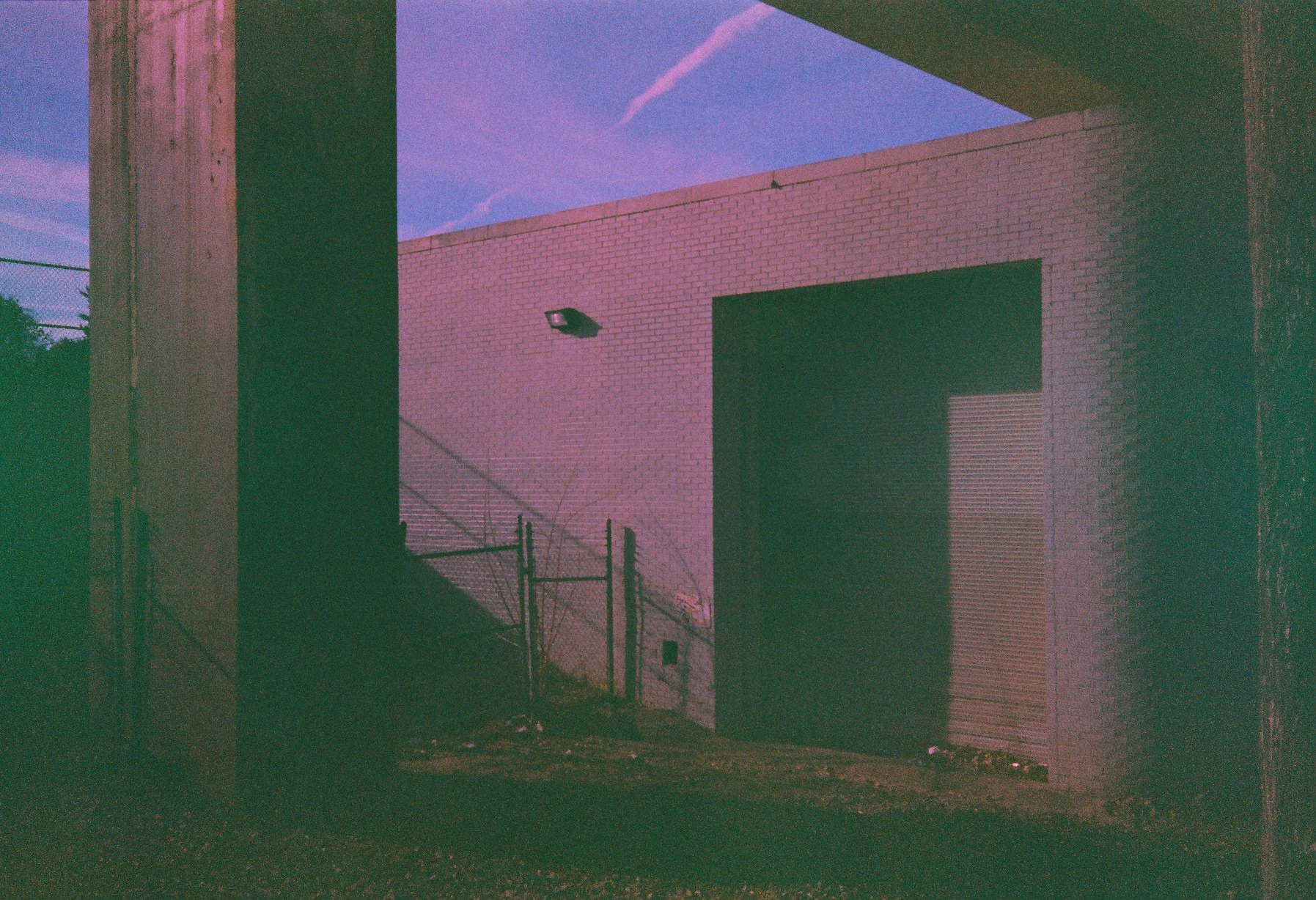 Nighttime view of a building with brick walls and a large black door, with a chain-link fence and light fixture in foreground, purple and blue sky in background.